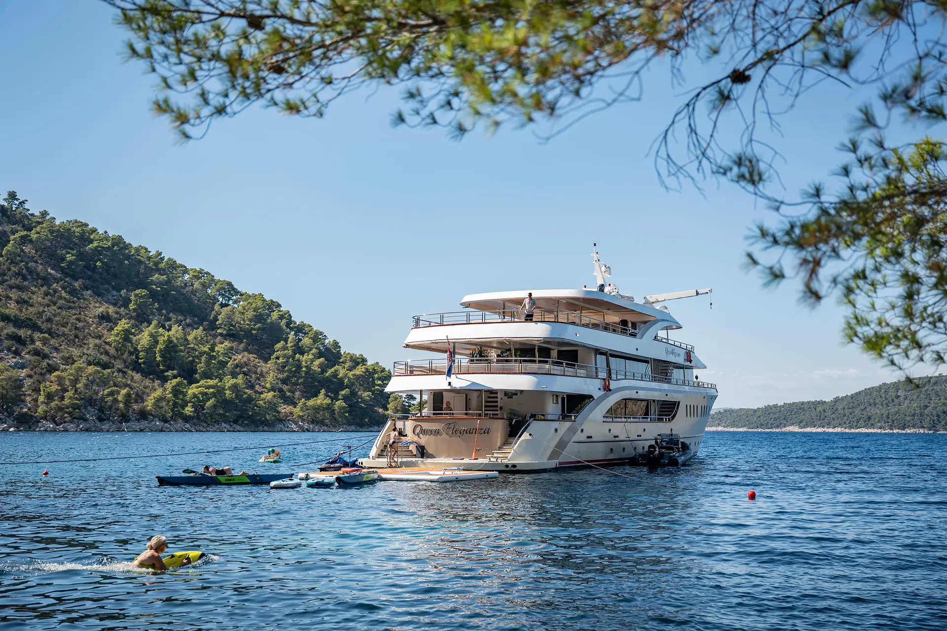 A large white yacht named Queen Eleganza is anchored near a forested shoreline, with people kayaking and swimming in the clear blue water under a sunny sky. Pine branches frame the top of the image.