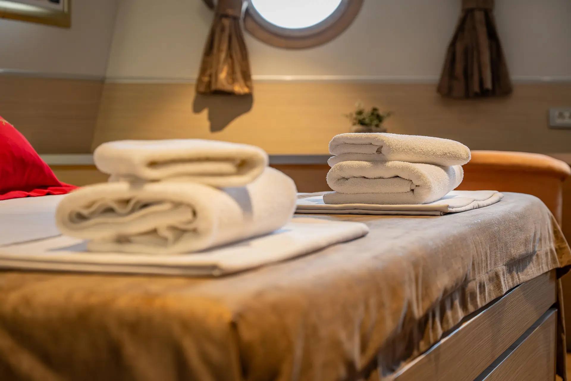 Two neatly folded white towels rest on a bed in a warmly lit room, with a circular window and tan curtains in the background.