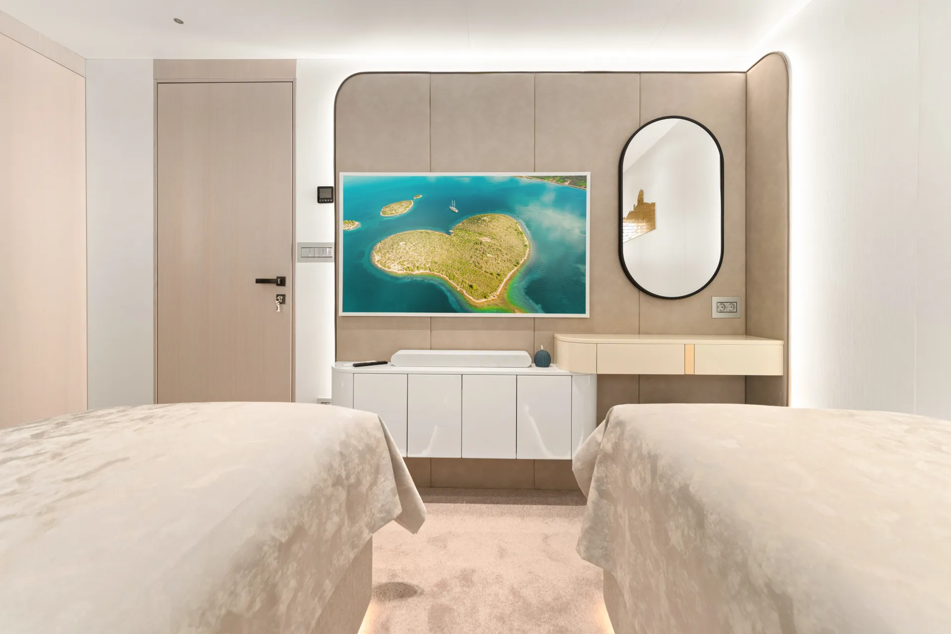 MAXITA A modern, minimalist bedroom with two beds, a wall-mounted TV displaying an island scene, a round-edged mirror, a sleek console table, and neutral-toned decor.