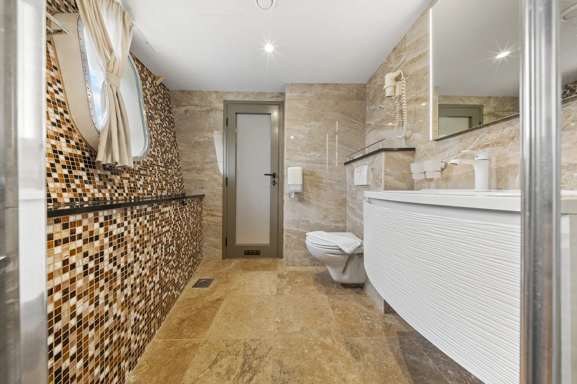 Modern bathroom with tan stone tile, mosaic accent wall, round window with beige curtains, floating toilet, curved white vanity, large mirror, and wall-mounted hairdryer. Brightly lit with a clean, contemporary design.