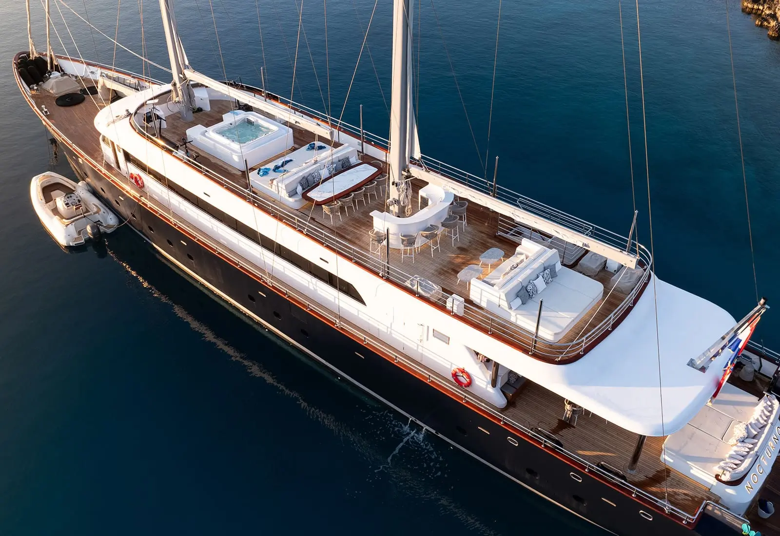 Aerial view of a luxury yacht on calm water, featuring a hot tub, lounge chairs, dining area, and sunbathing spaces on the deck, with a small boat moored alongside.