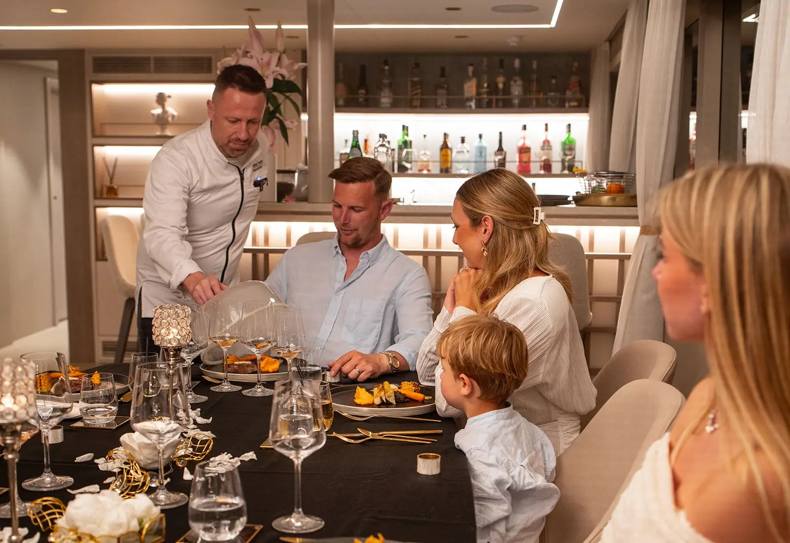 A chef serves food to a seated man at an elegant dining table with three women and a child. The modern room features a well-lit bar in the background.