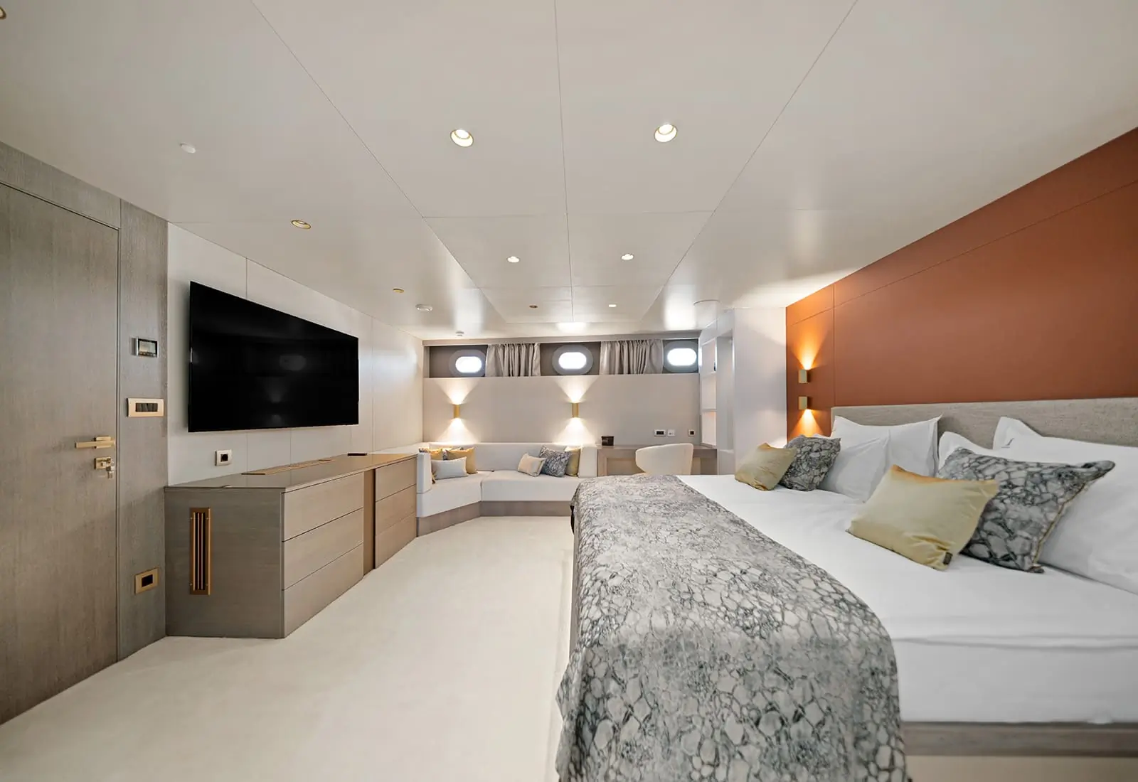 Modern yacht bedroom with a large bed, patterned bedding, pillows, wall-mounted TV, built-in cabinets, small sofa, and porthole windows. Neutral tones and soft lighting create a luxurious, spacious feel.