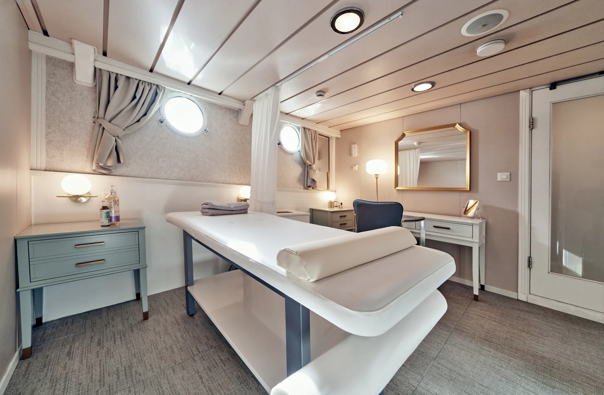 LA PERLA A bright, modern spa treatment room with a padded massage table in the center, two side tables with lamps, towels, and a vanity with a mirror. The room has porthole windows and soft, neutral decor.