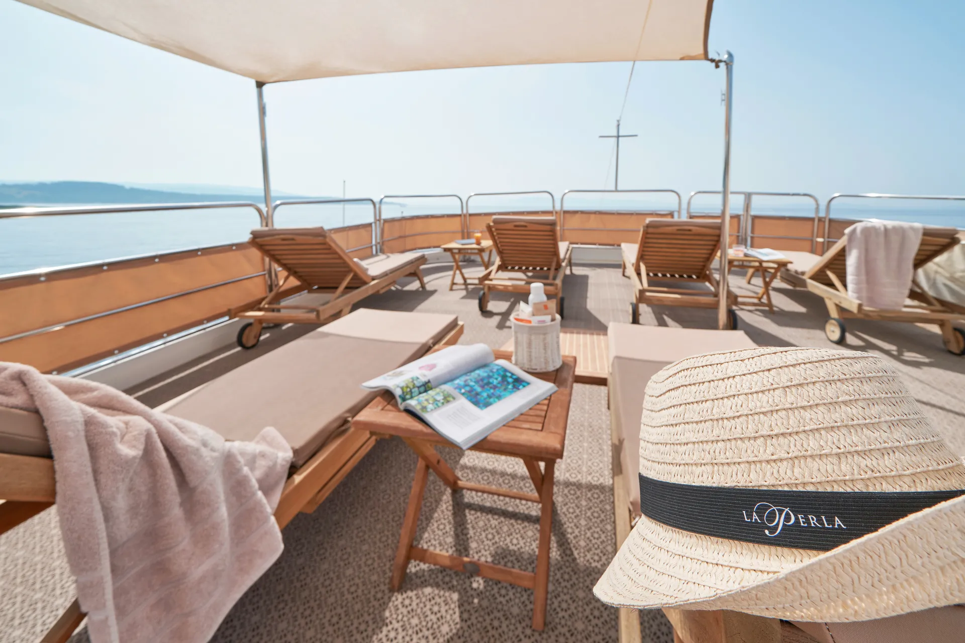 LA PERLA Sun loungers with towels on a shaded deck overlooking the sea, a wooden table with books, sunscreen, and a straw hat labeled “La Perla” in the foreground, creating a relaxing atmosphere.
