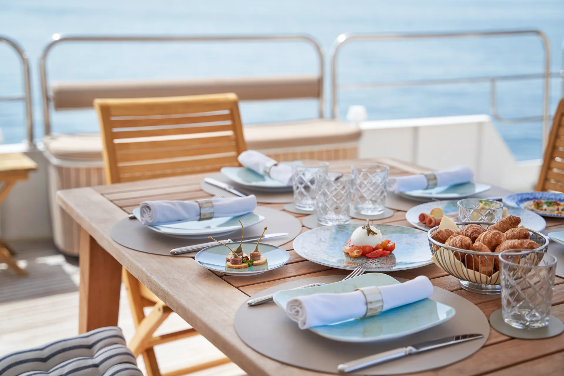 LA PERLA A wooden table set for a meal on a yacht deck, overlooking the sea. Plates, cutlery, napkins, glasses, and a variety of breakfast foods, including croissants and fruit, are neatly arranged in the sunlight.