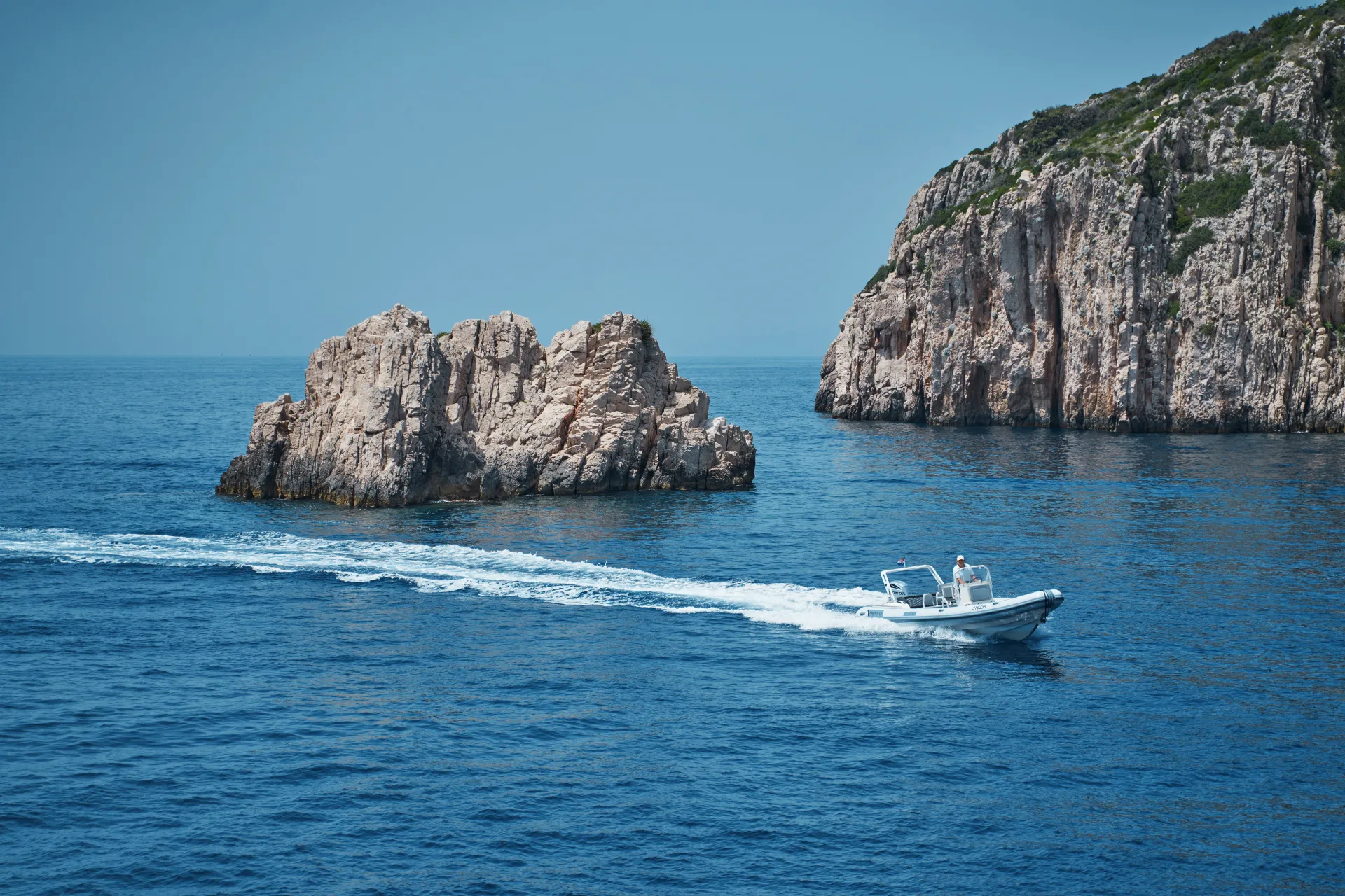 LA PERLA A small white motorboat moves quickly across clear blue water, leaving a trail of white foam behind. Rocky outcrops covered in greenery rise from the sea under a bright, clear sky.