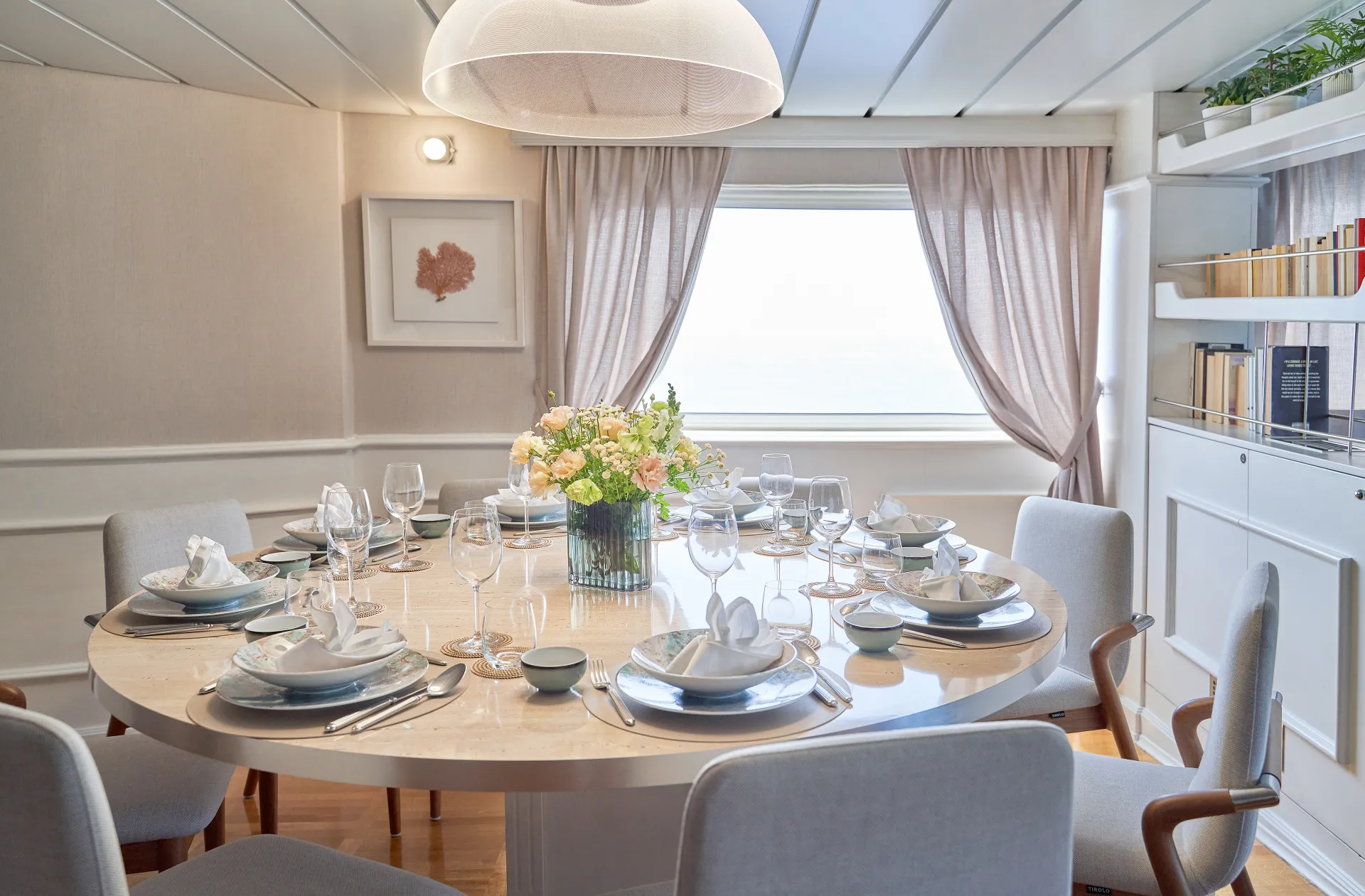 LA PERLA A round dining table set for eight with elegant dishware, glassware, and a floral centerpiece, surrounded by cushioned chairs in a bright room with a large window and light curtains. Bookshelves and wall art are in the background.