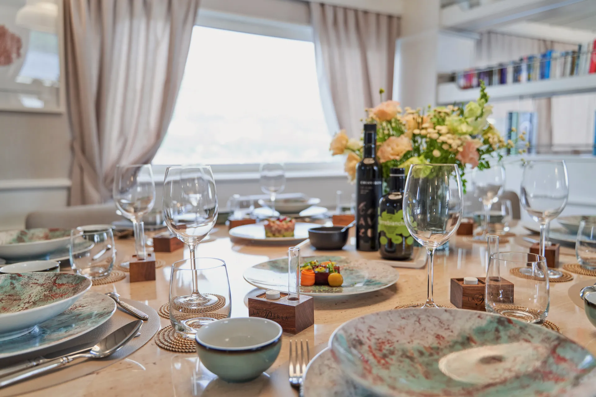 LA PERLA Elegant dining table set with artistic plates, wine glasses, and floral centerpiece. Bottles of wine and small gourmet dishes are arranged neatly, with soft natural light coming through a large window in the background.