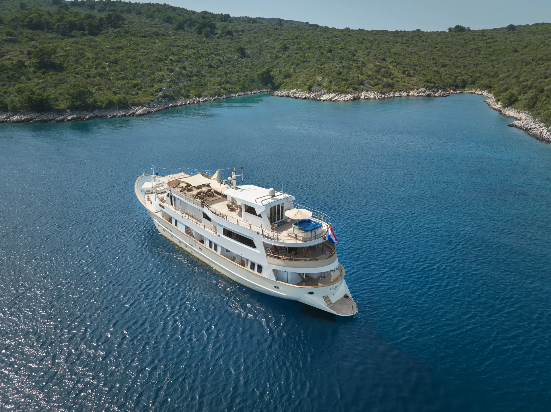 LA PERLA A large white yacht with multiple decks sails in calm blue water near a lush, green coastline under a clear sky.