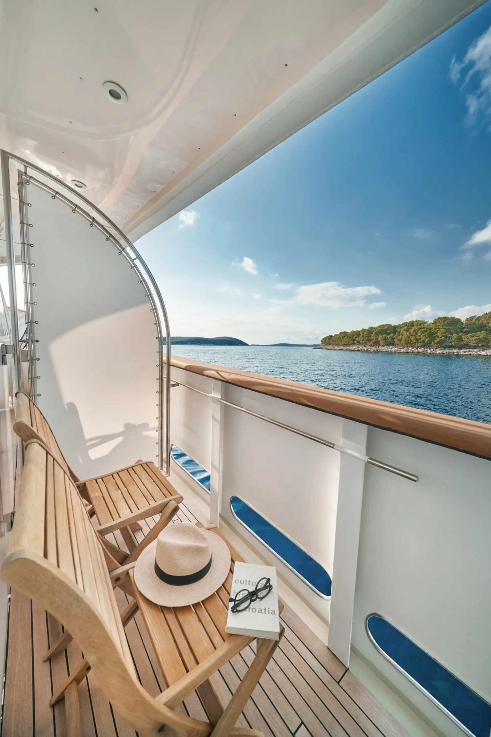 LA PERLA A wooden bench and table on a yacht deck overlook calm blue water and a distant tree-covered shore; a hat and a book titled Croatia rest on the bench under a sunny sky.