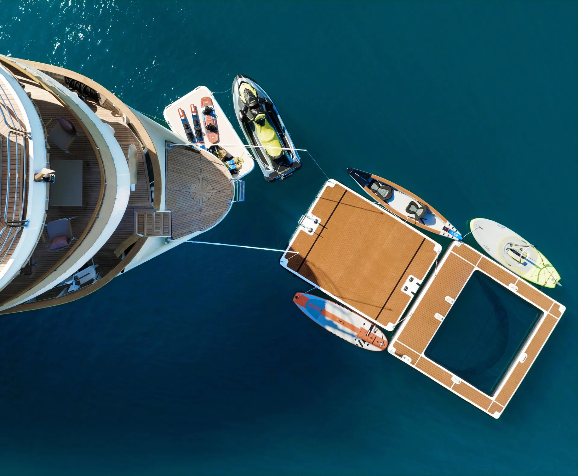 LA PERLA Aerial view of a yacht anchored in clear blue water with floating platforms, jet skis, paddleboards, and small boats tied alongside.