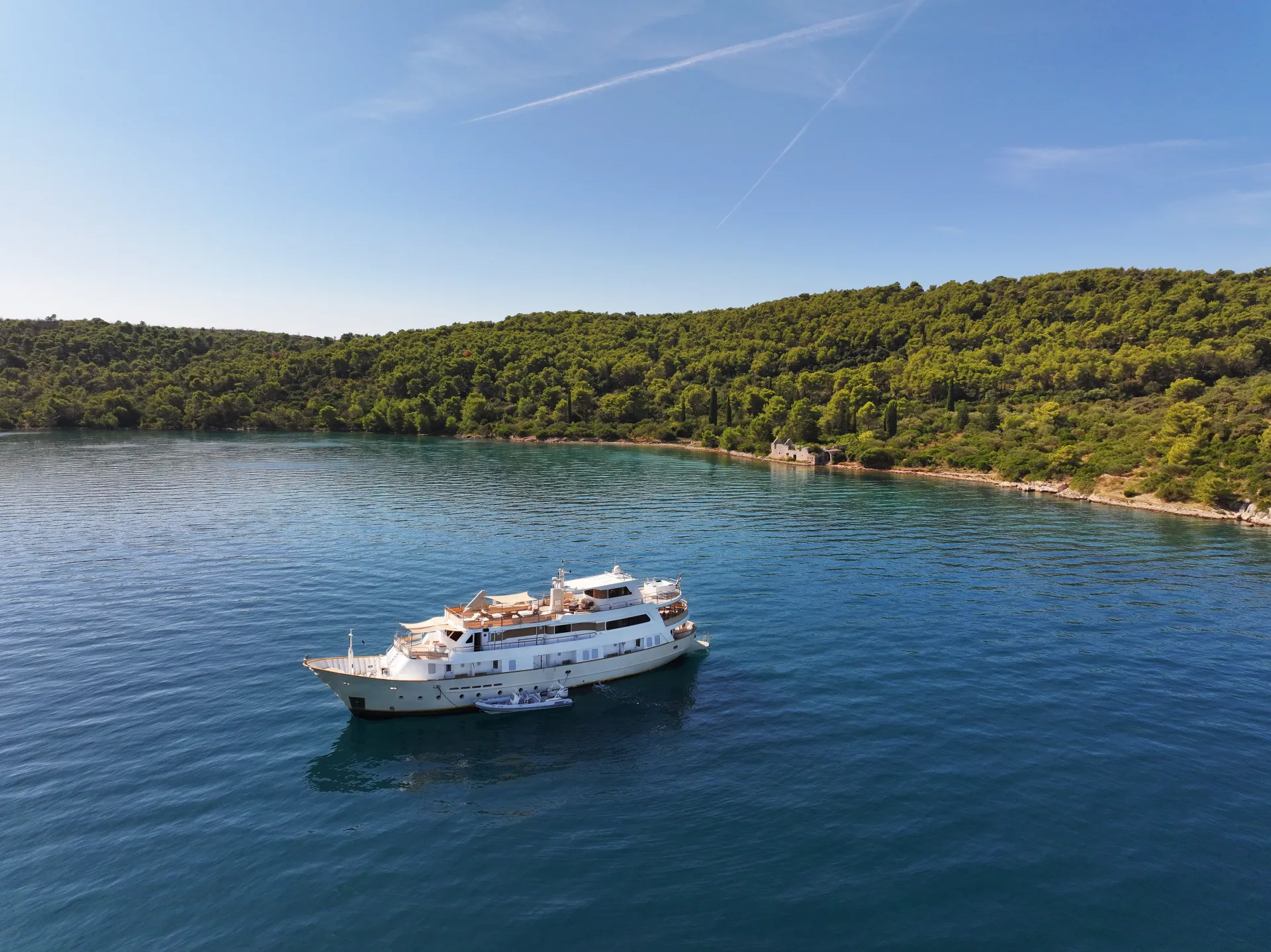 LA PERLA A white yacht floats on calm blue water near a lush green forested shoreline under a clear, sunny sky.
