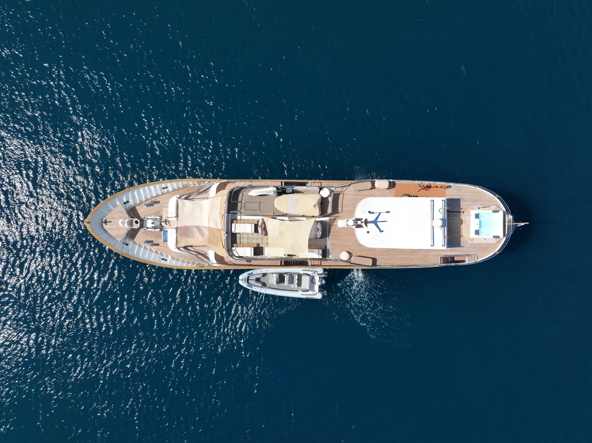 LA PERLA Aerial view of a large luxury yacht anchored on calm, deep blue water, with a smaller boat tied alongside. The yacht has decks with lounge areas and a swimming pool at the rear.
