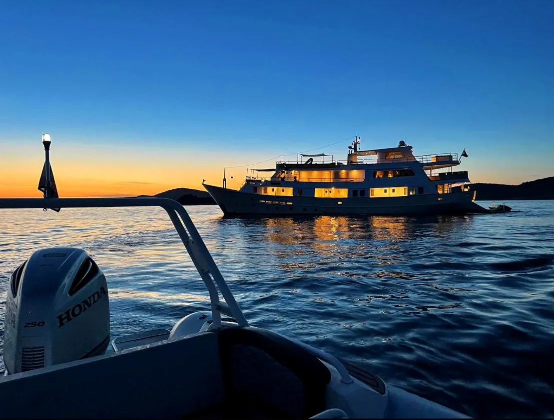 LA PERLA A large, well-lit yacht is anchored on calm water at sunset, with reflections on the surface. In the foreground, part of a smaller boat with a Honda motor is visible. The sky is deep blue with an orange glow at the horizon.