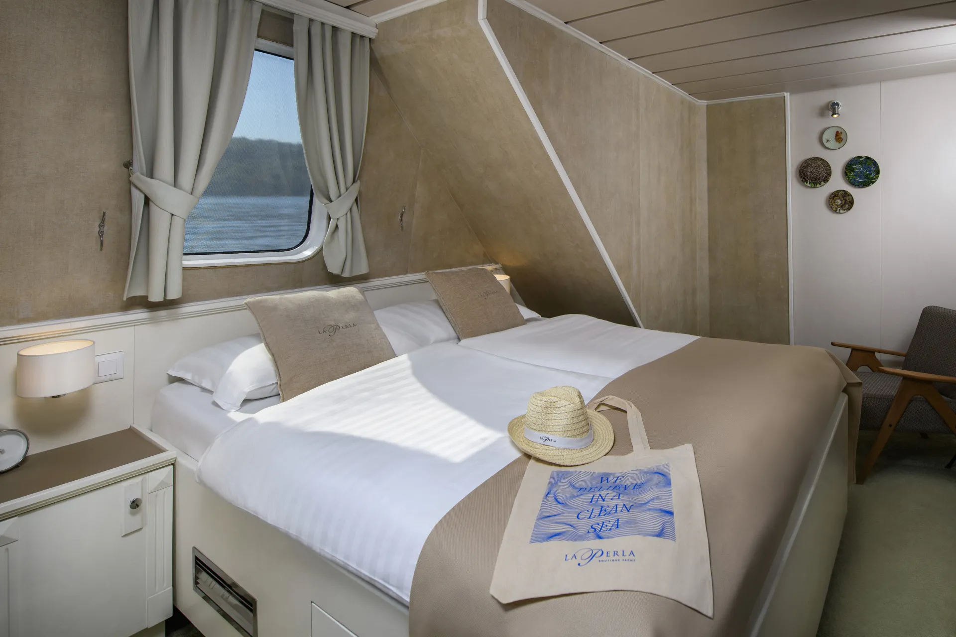 LA PERLA A cozy cruise ship cabin with a large neatly made bed, beige bedding, a window with curtains, and a nightstand. A straw hat and tote bag rest on the bed. Wall art and a chair are visible in the background.