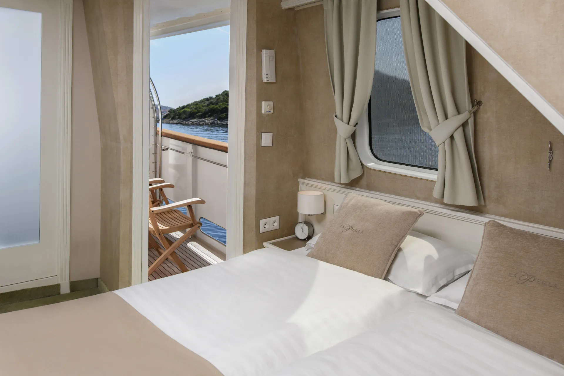 LA PERLA A cozy bedroom with a double bed, beige walls, and light brown pillows, opens onto a deck with two wooden chairs overlooking water and greenery outside the window.