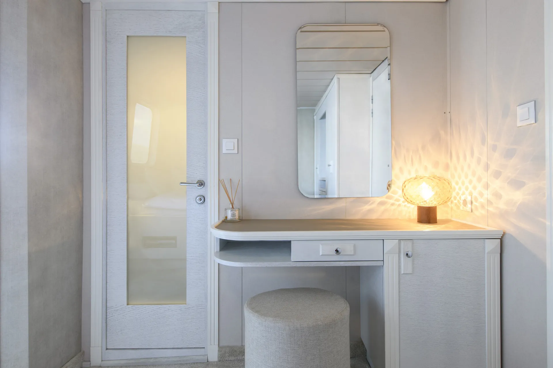 LA PERLA A minimalistic vanity with a round cushioned stool, a rectangular mirror, and a small lamp casting patterned light sits next to a door with a frosted glass panel in a light-colored room.