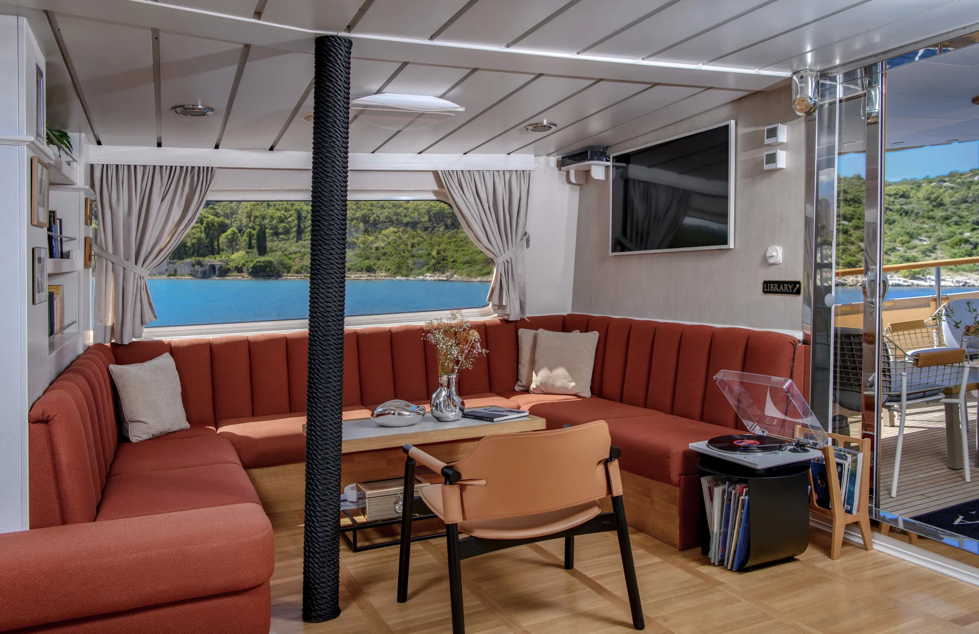 LA PERLA A cozy yacht lounge with orange L-shaped sofas, a table with flowers and books, a TV on the wall, large windows with open curtains, and a view of blue water and green hills outside.