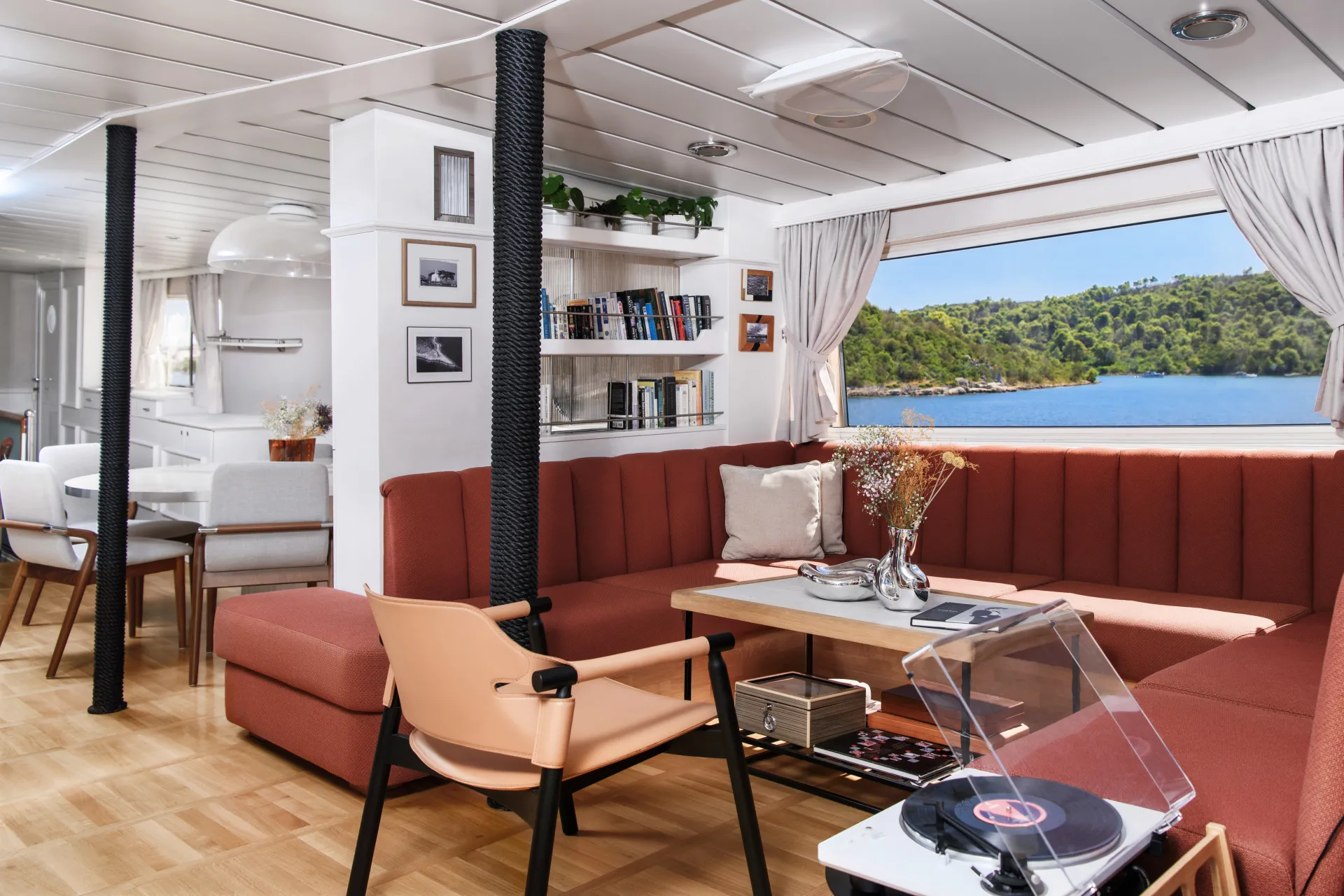 LA PERLA A modern, cozy lounge with rust-colored sofas, a record player, bookshelves, and large windows showing a scenic lake and green hills. The space has wood floors and white walls, with a dining area in the background.