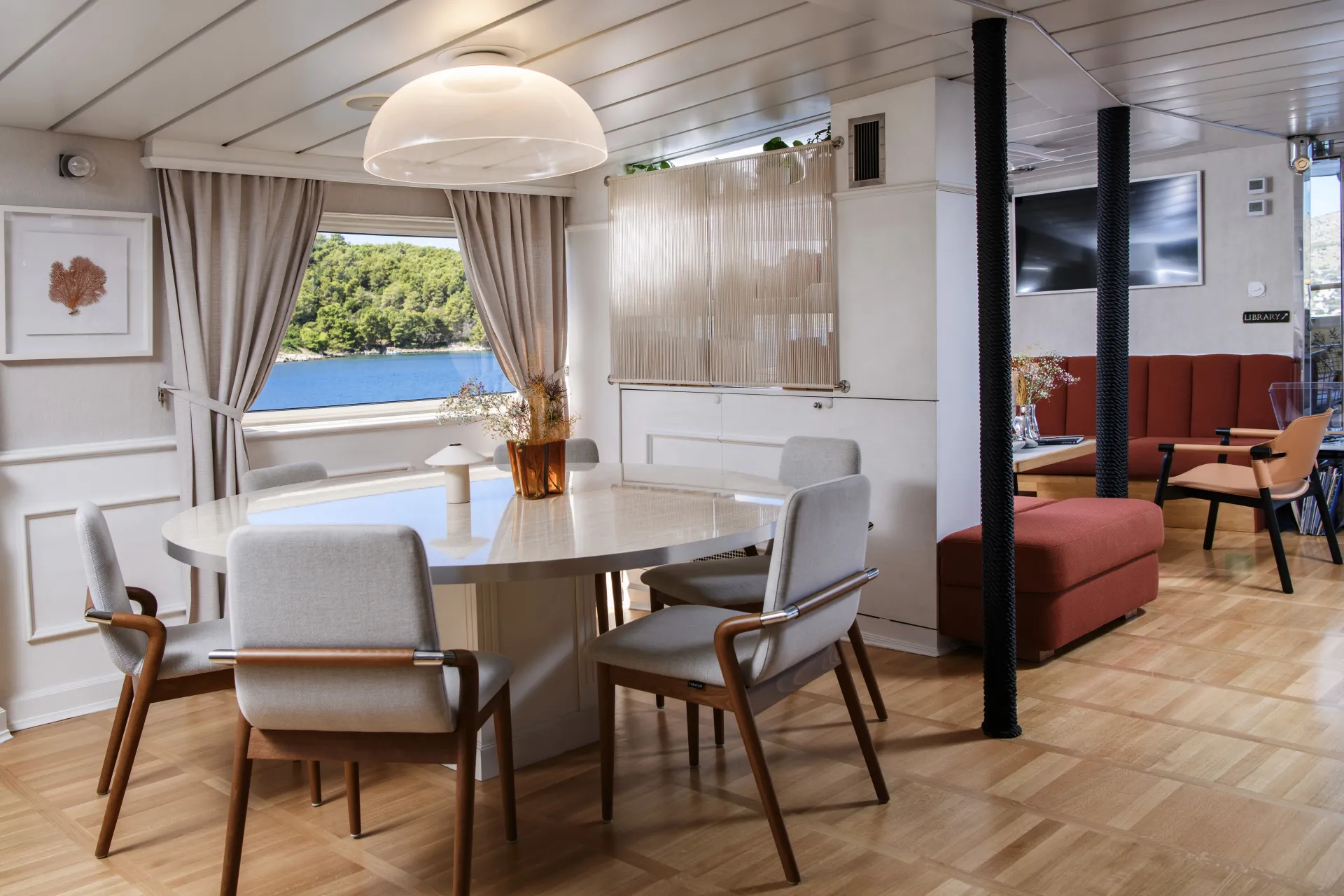 LA PERLA Modern dining area with a round table and six cushioned chairs, set near a window with a view of water and trees outside. Neutral tones and wood flooring create a cozy, inviting atmosphere.