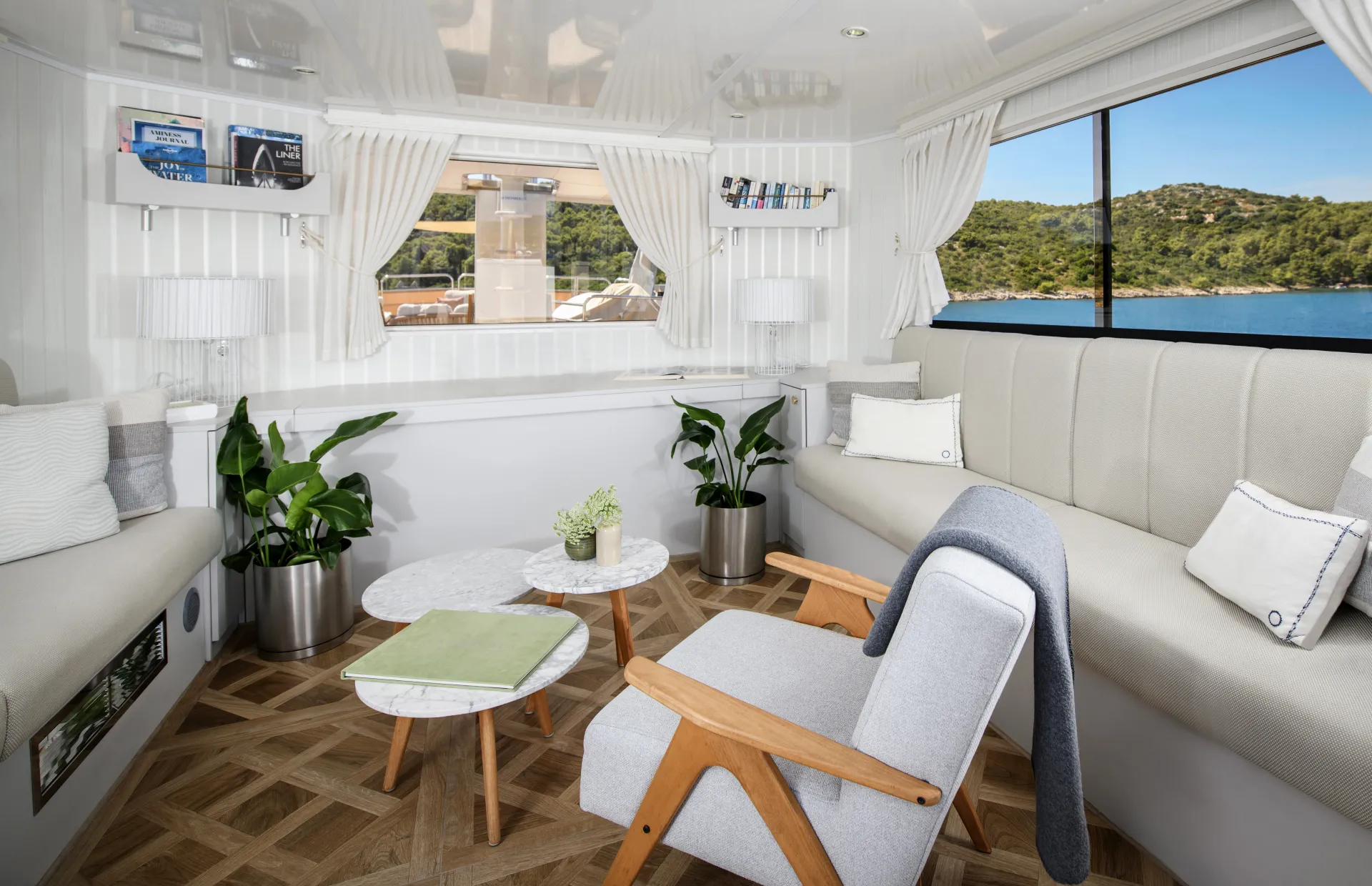 LA PERLA Bright, modern living space with light wood floors, neutral sofas, an armchair, round marble tables, potted plants, shelves with books, large window with a view of green hills and water, and white curtains.