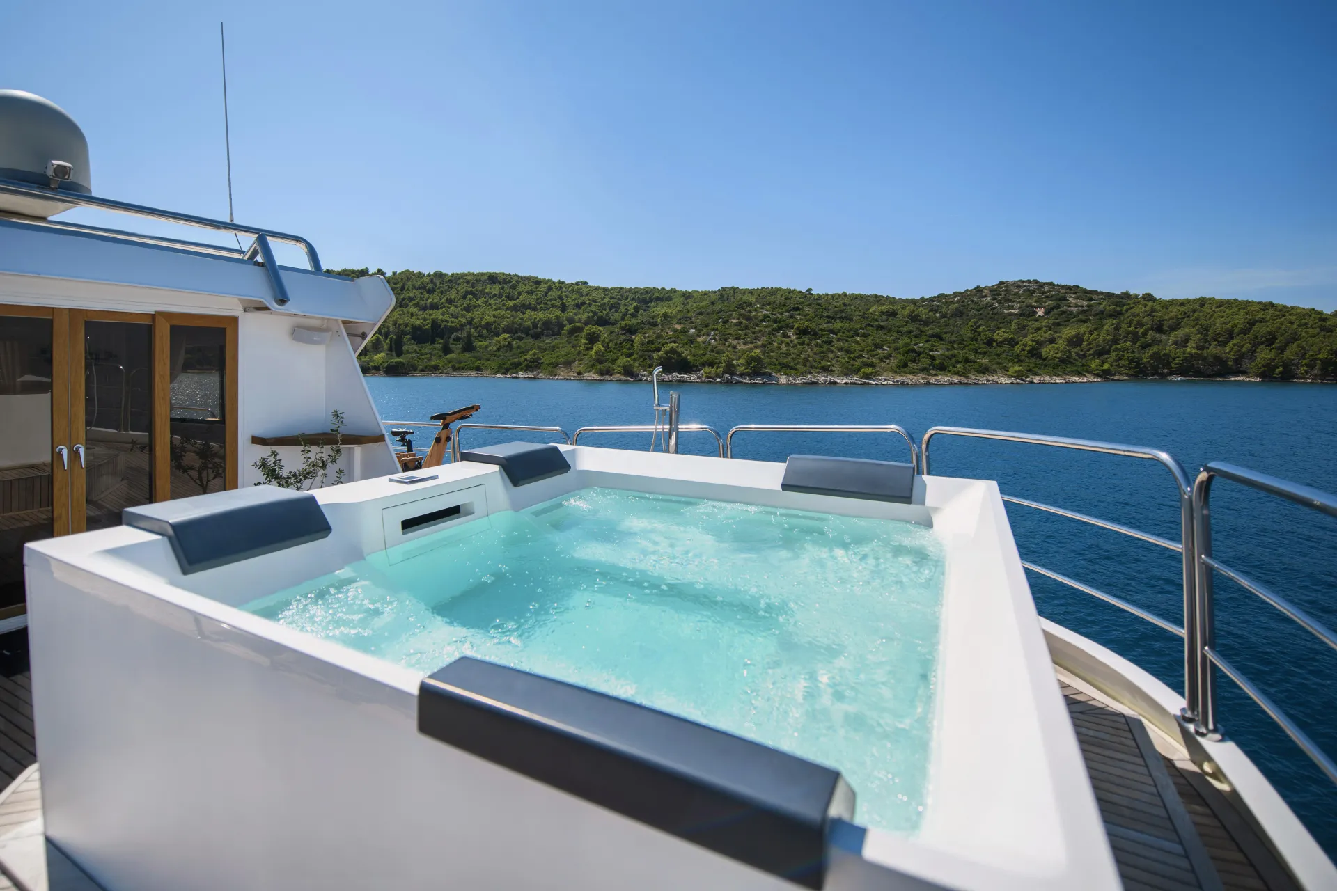 LA PERLA A luxury yacht features an outdoor hot tub with bubbling water, surrounded by metal railings. The yacht is anchored near a green, forested shoreline under a clear blue sky.