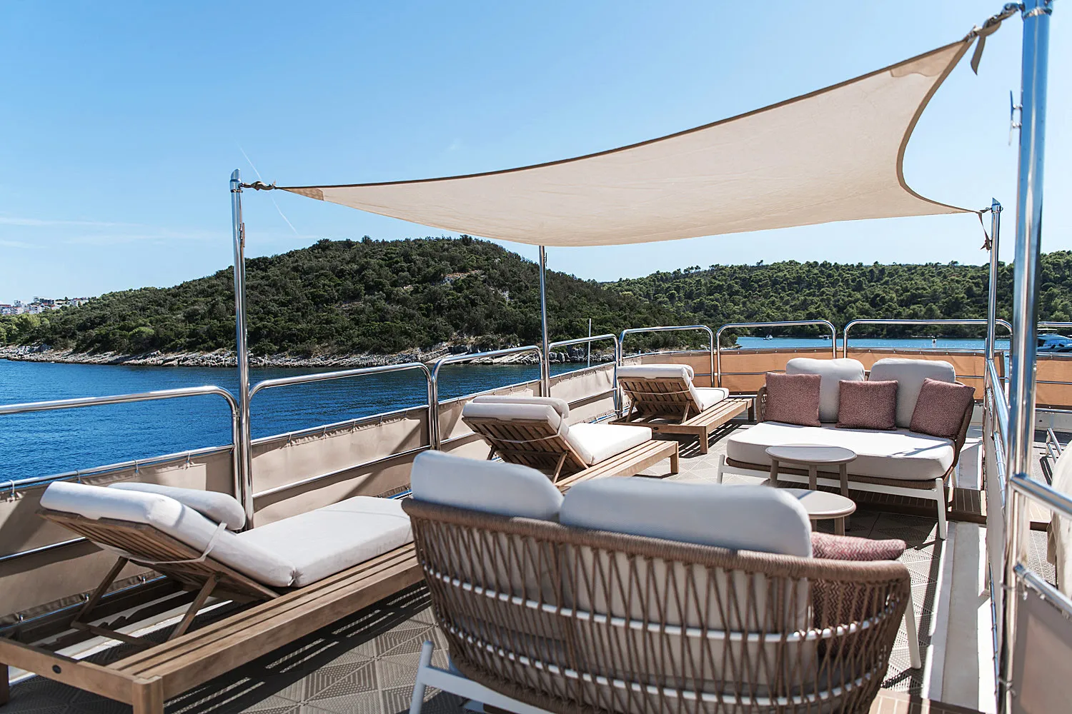 LA PERLA A yacht deck with cushioned lounge chairs and sofas under a sunshade, overlooking calm blue water and a green, hilly shoreline on a sunny day.