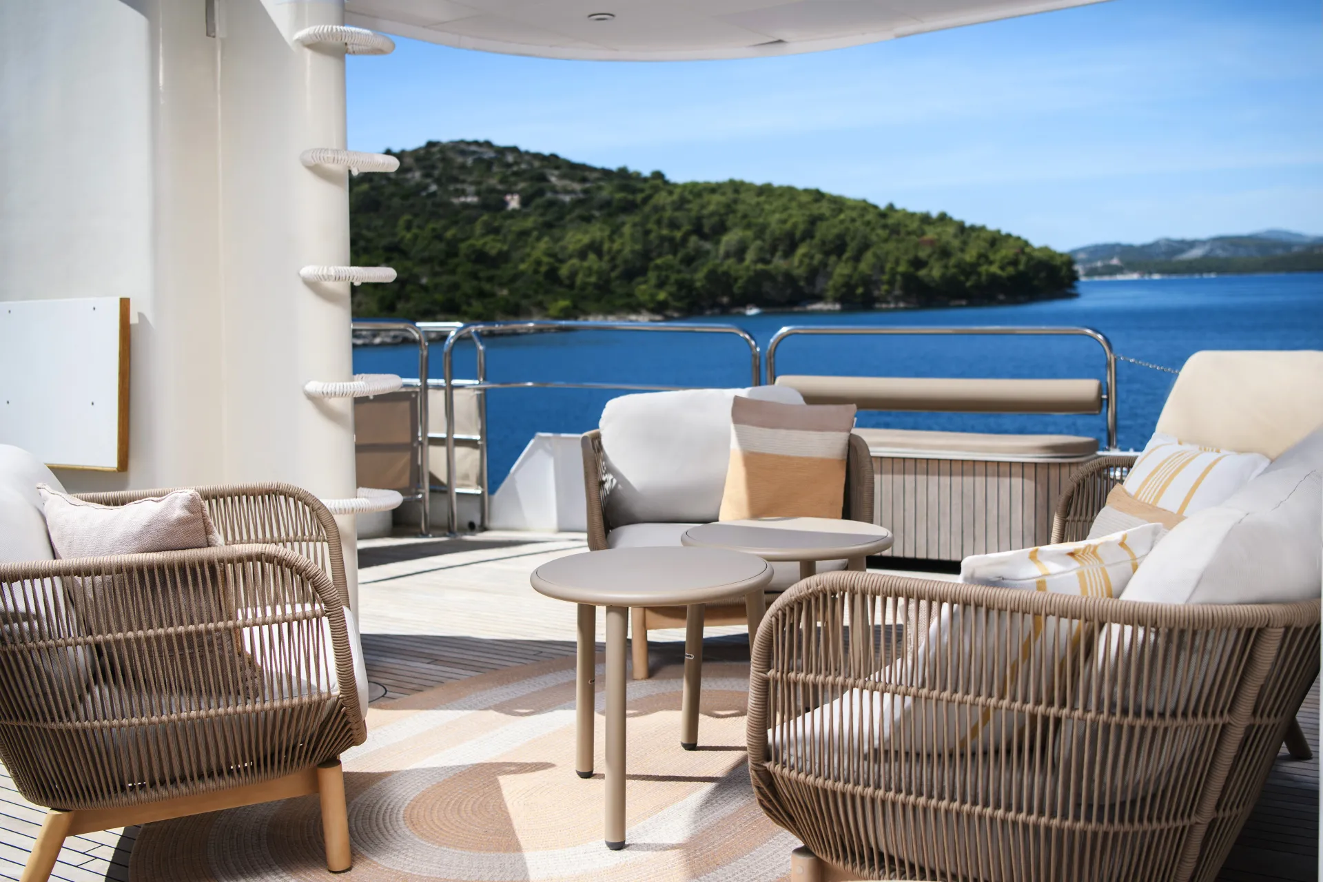 LA PERLA Outdoor seating area on a yacht with wicker chairs, a table, and cushions, overlooking blue water and a forested coastline under a clear sky.