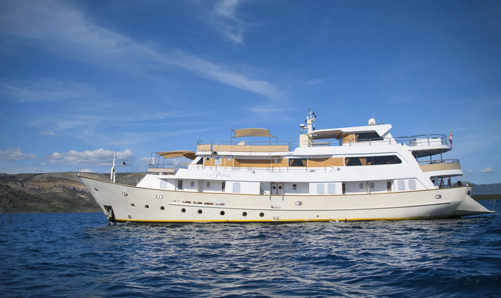 LA PERLA A large white luxury yacht with multiple decks floats on calm blue water under a clear sky, with a distant shoreline visible in the background.