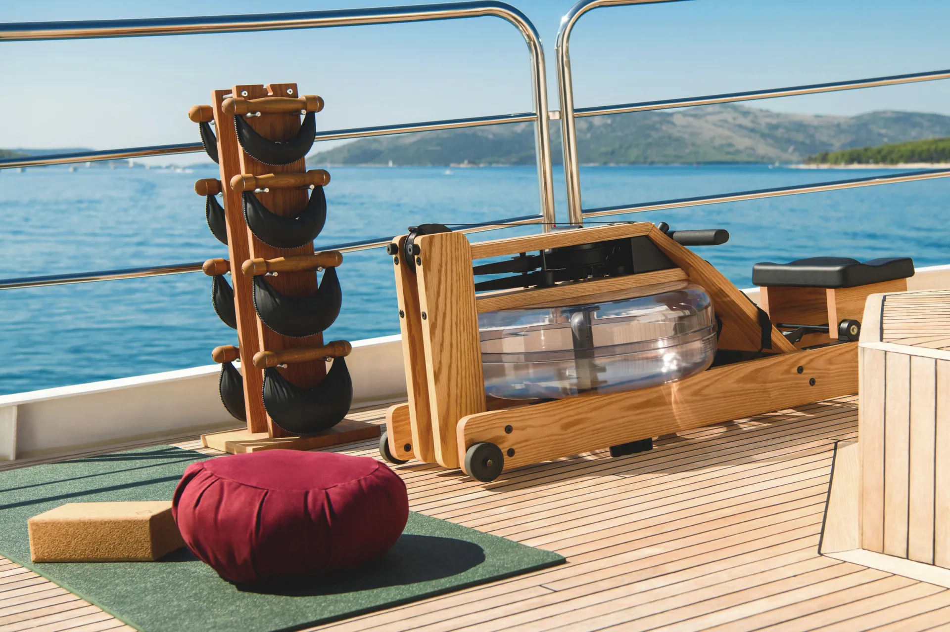LA PERLA A wooden rowing machine, black kettlebells on a rack, a red cushion, and yoga blocks are set on a yacht deck with a scenic view of blue water and distant hills.