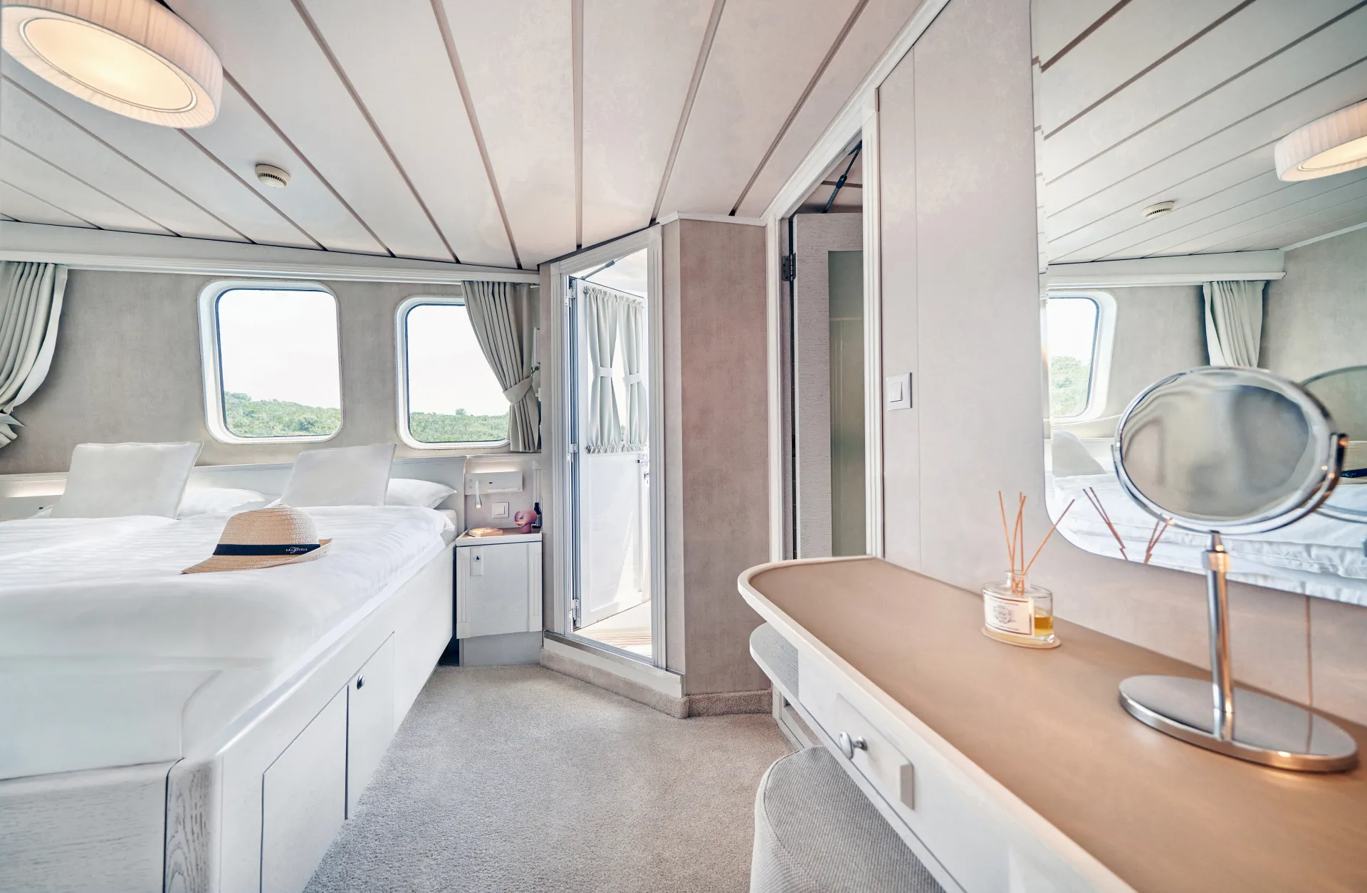 LA PERLA A bright, modern bedroom with large windows, white walls, and a neatly made bed. There’s a hat on the bed, a vanity with a mirror, and an open door leading to a small balcony with outdoor views.