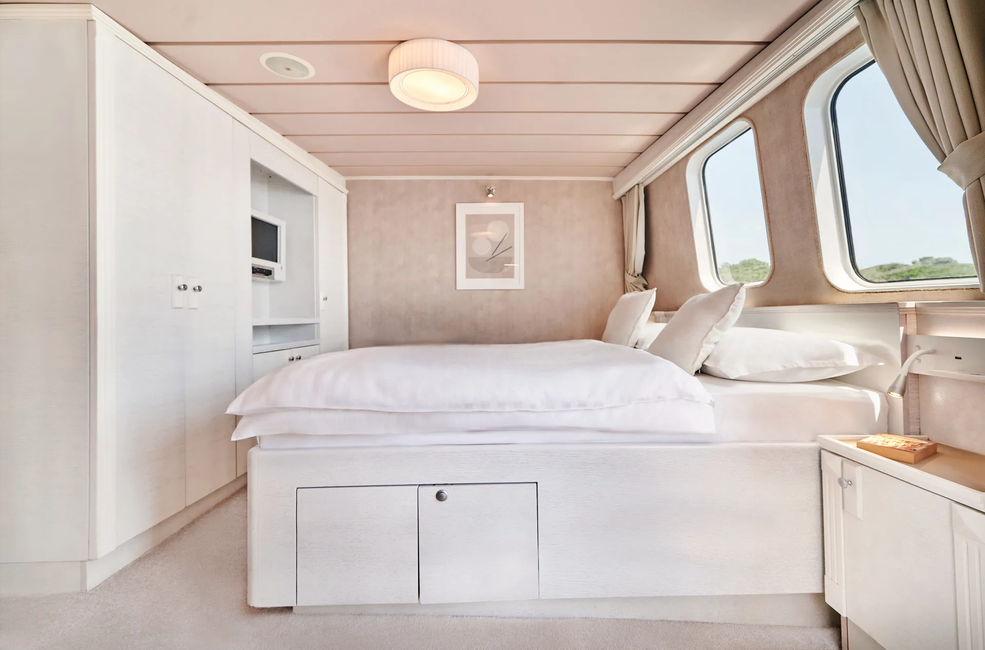 LA PERLA A bright, modern bedroom with a neatly made white bed, built-in storage, a small TV, large windows with curtains, and soft natural light illuminating the space.