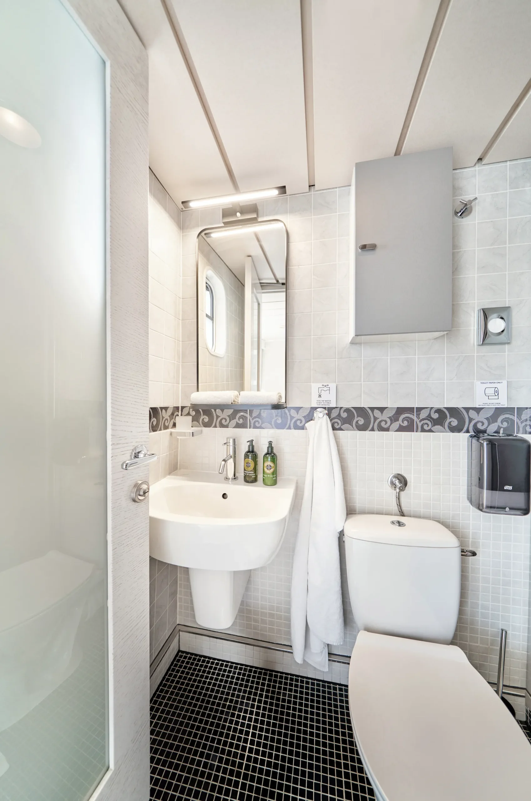 LA PERLA Modern bathroom with white tiles, a wall-mounted sink, and a toilet. A mirror, cabinet, towel, soap dispenser, and toiletries are visible. The floor has small black tiles and natural light enters through a frosted glass door.