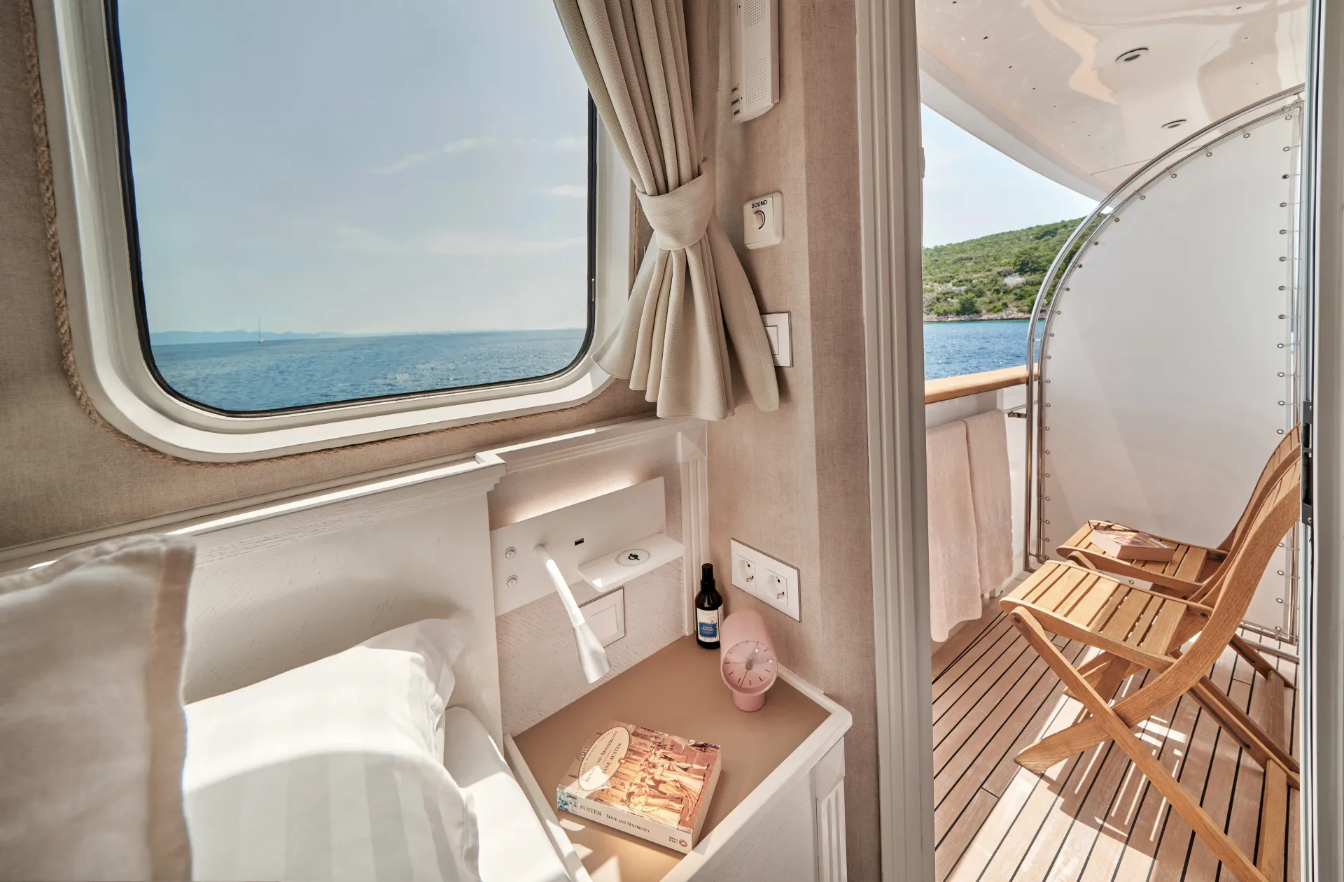 LA PERLA A cozy cabin with a made bed, book, and drink on the nightstand overlooks a sunny private balcony with two wooden chairs and a sea view through large windows and an open door.