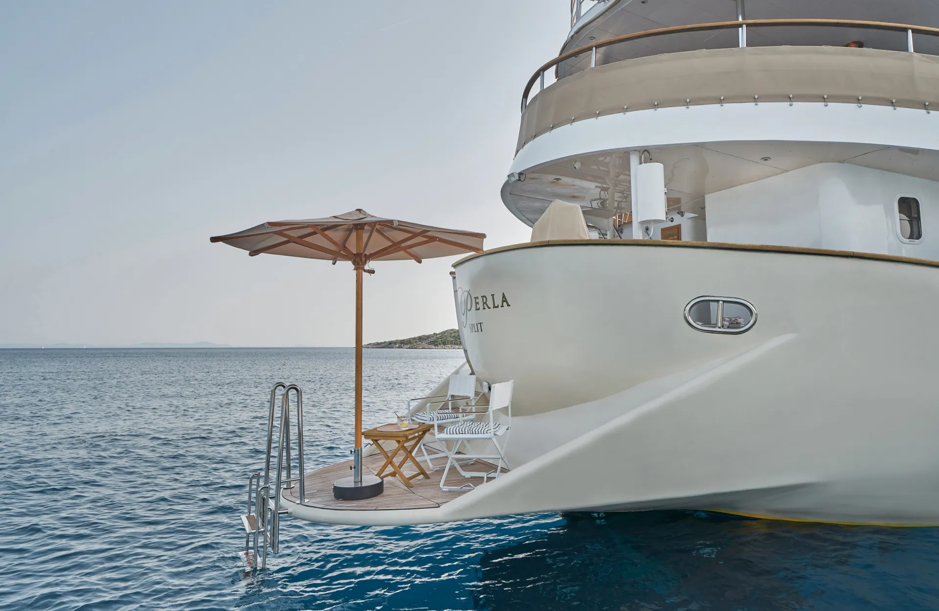 LA PERLA The rear deck of a luxury yacht with a sun umbrella, chairs, table, and a ladder leading into calm blue sea, with clear skies in the background.