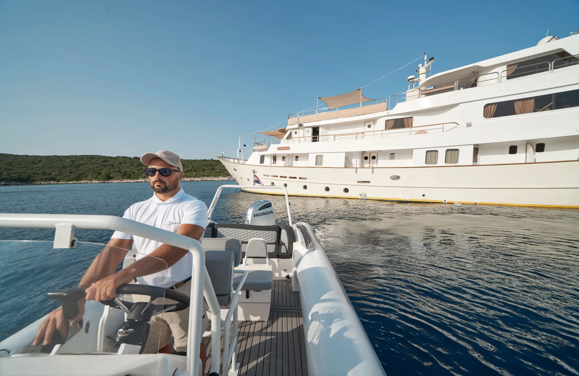 LA PERLA A man wearing sunglasses and a cap steers a small inflatable boat on calm water, with a large white yacht and green shoreline in the background under a clear blue sky.