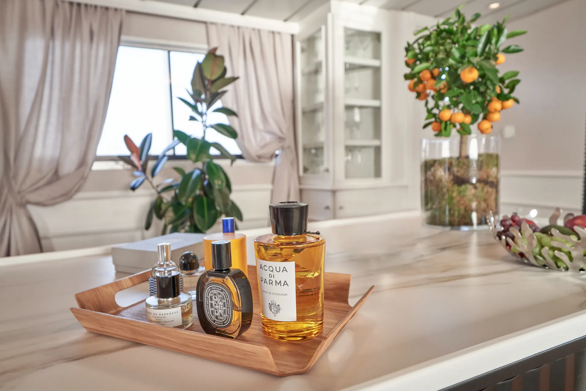 LA PERLA A wooden tray holding several luxury perfume bottles sits on a marble countertop. In the background are potted plants, including a small citrus tree with orange fruit, and sunlight streams through draped curtains.