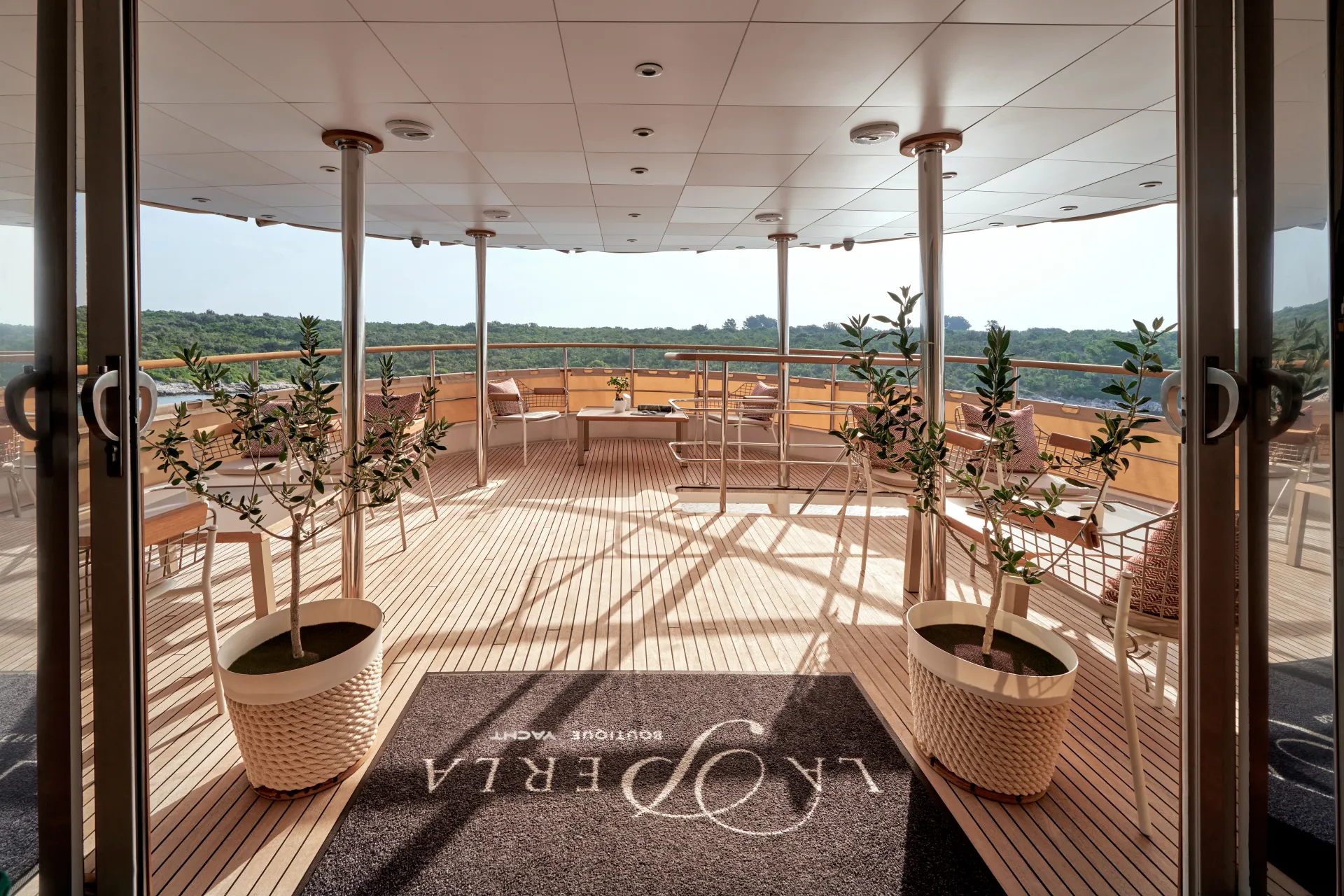 LA PERLA Spacious yacht deck with potted plants, glass railings, wood flooring, and patio furniture. There is a doormat labeled LYPERA BOUTIQUE YACHT and a view of green trees beyond the deck.