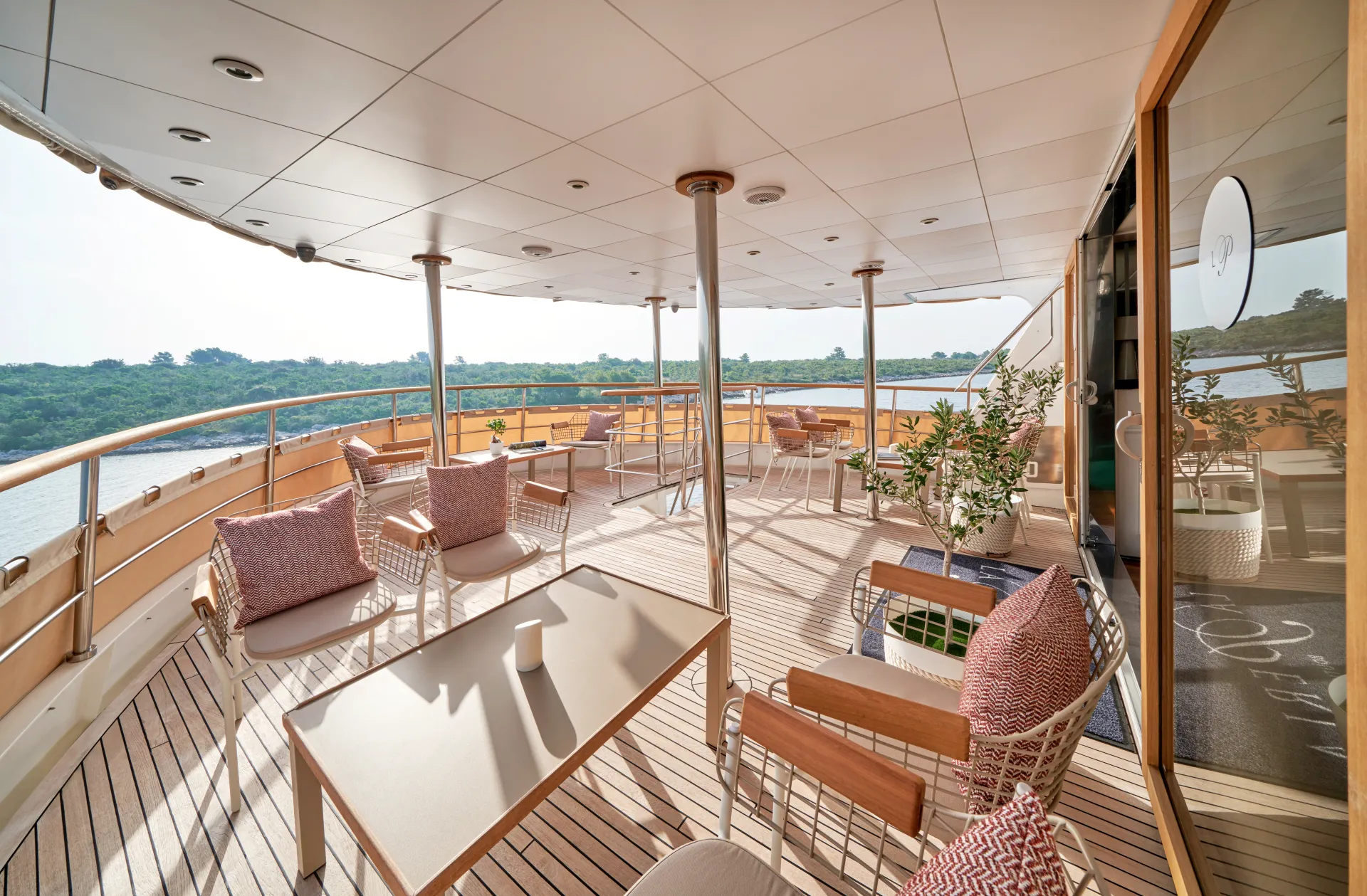 LA PERLA Spacious yacht deck with modern lounge chairs, tables, and potted plants, surrounded by railings and overlooking a river and lush greenery.