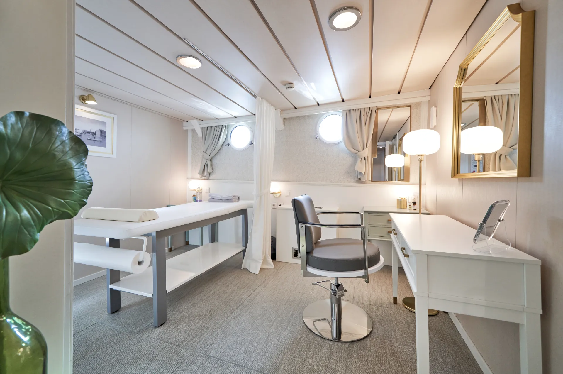 LA PERLA A bright, modern spa room with a massage table, a salon chair at a vanity, mirrors, lamps, and two porthole windows. The decor is neutral with white furniture and light wood accents.