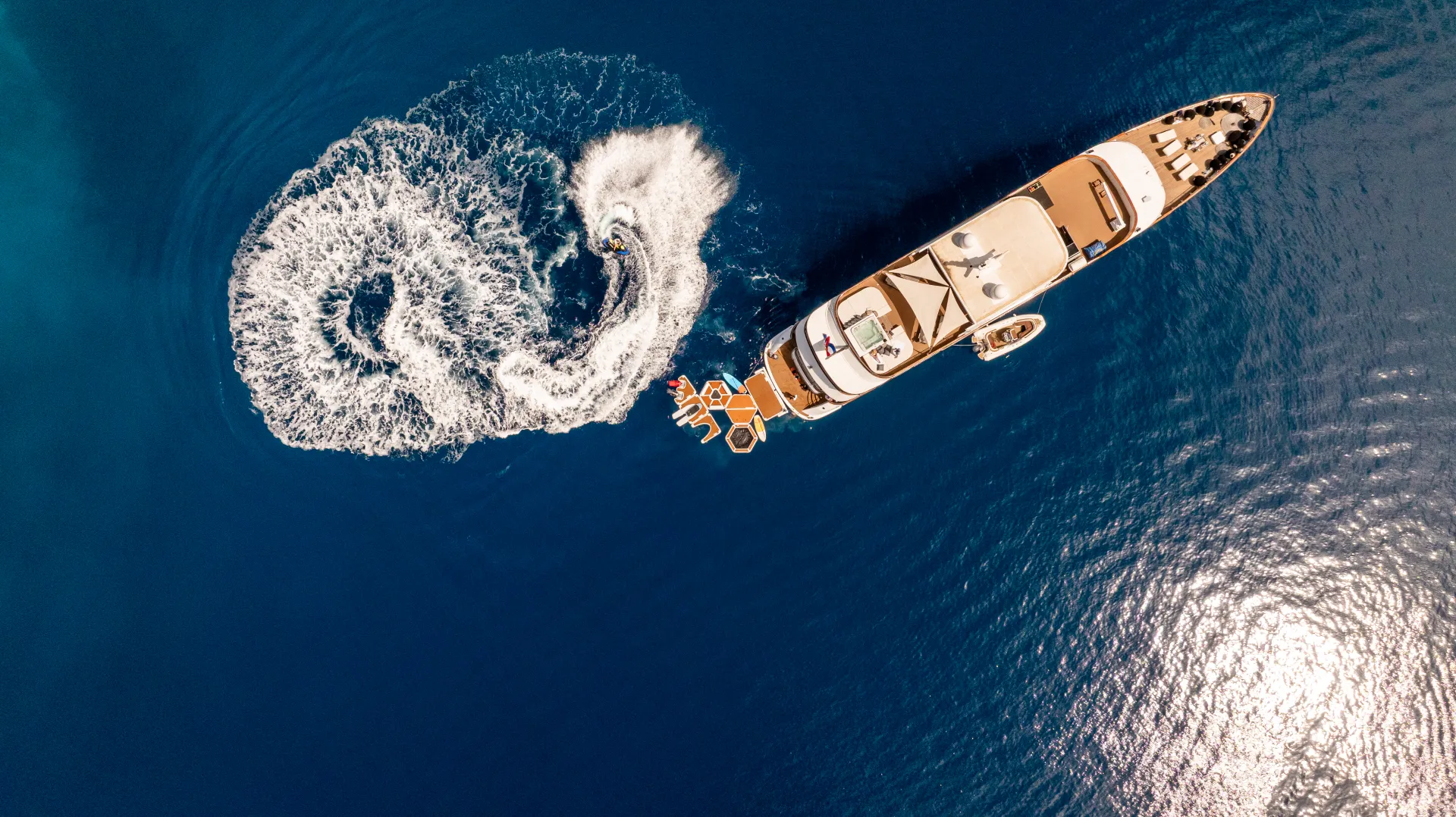 CRISTAL Aerial view of a yacht on deep blue water, with a person on a jet ski creating circular waves beside it. Sunlight reflects off the water’s surface.