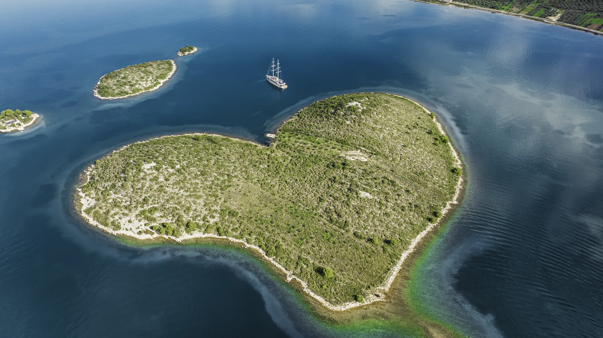 MAXITA Aerial view of a heart-shaped green island surrounded by blue water, with a boat sailing nearby and two smaller islands in the distance.