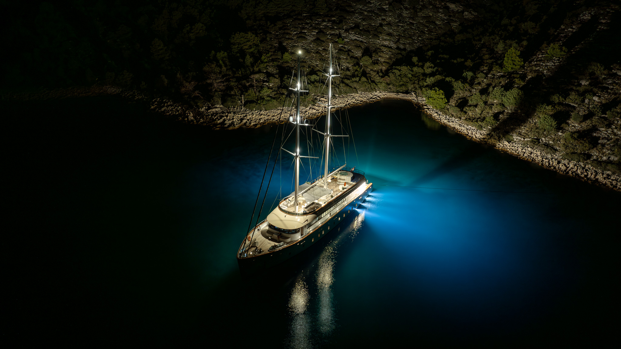 MAXITA A yacht anchored in a calm bay at night, illuminated by bright deck and underwater lights, surrounded by dark water and rocky, tree-lined shores.