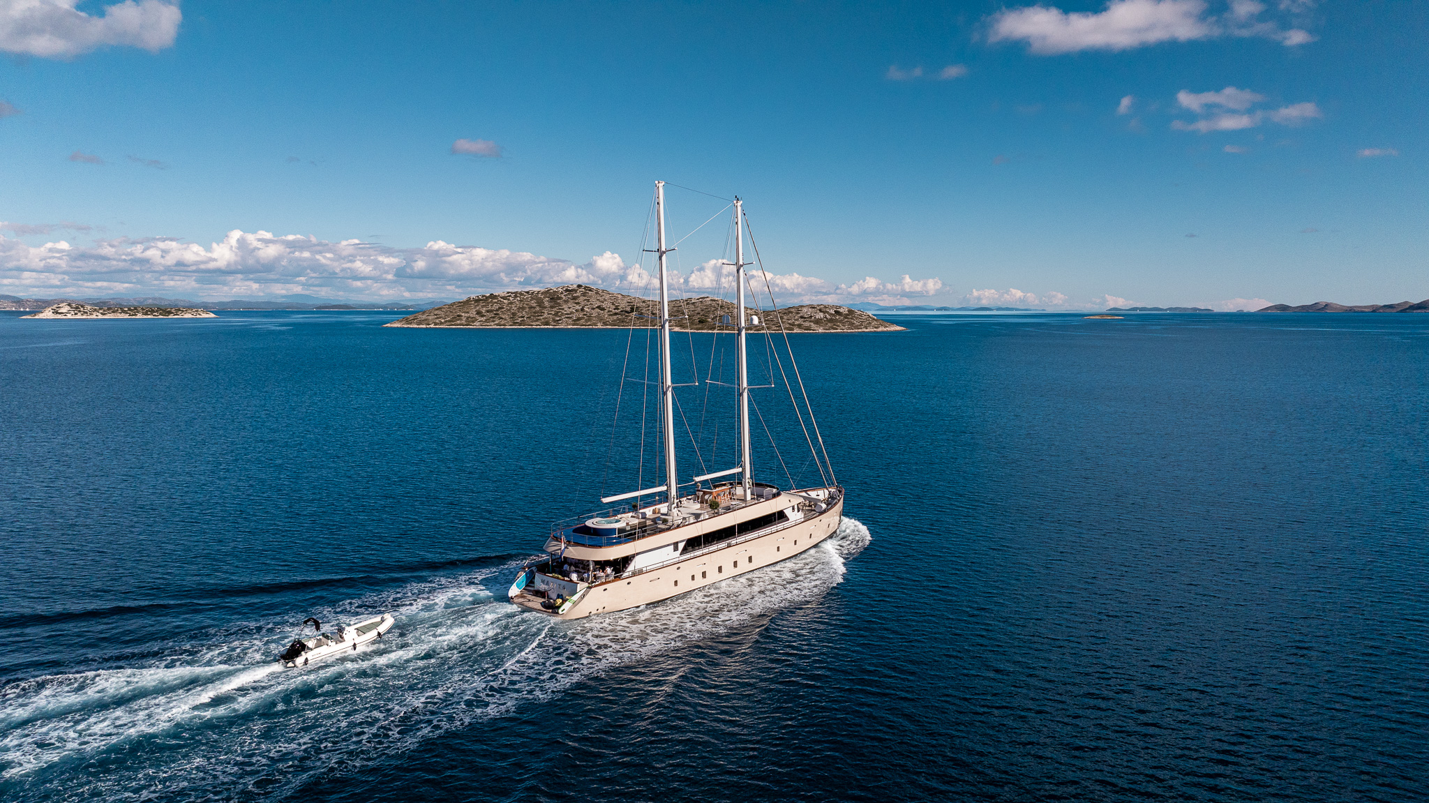 MAXITA A large sailboat yacht cruises on calm blue water, leaving a trail behind, with a small motorboat following. Small green islands and a blue sky with scattered clouds are visible in the background.