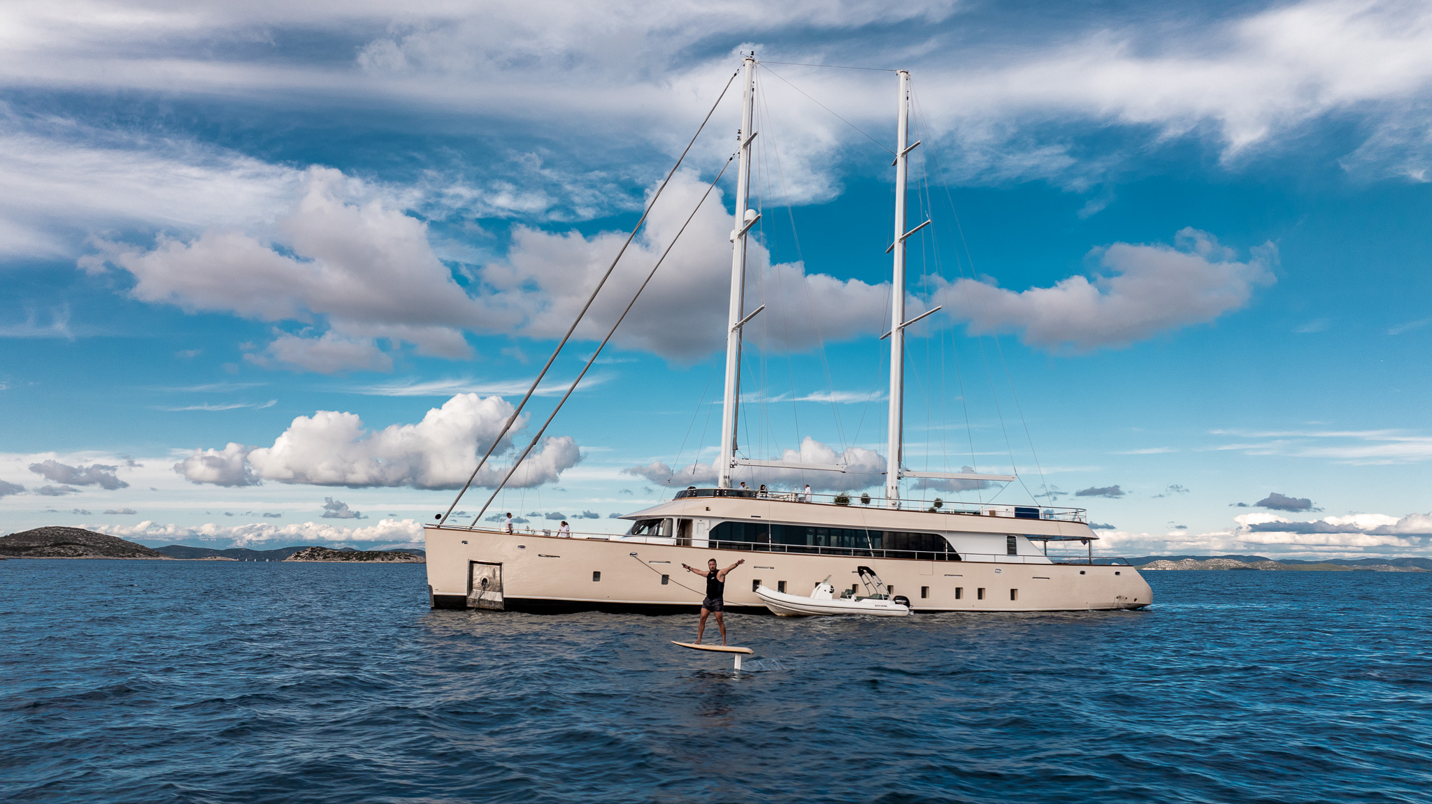 MAXITA A large, modern yacht floats on calm blue water under a partly cloudy sky. A person stands on a paddleboard nearby, arms raised in excitement. Small islands are visible in the distant background.