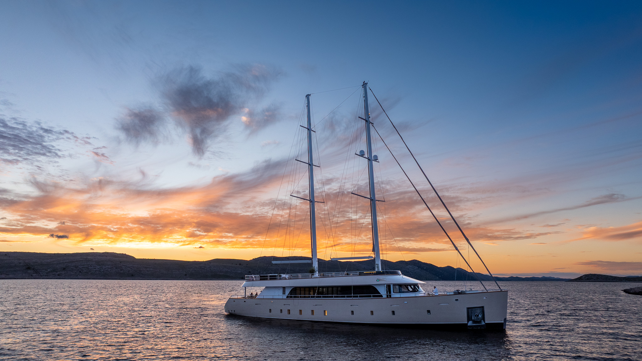 MAXITA A large white yacht with tall masts floats on calm water at sunset, with colorful clouds and distant hills in the background.