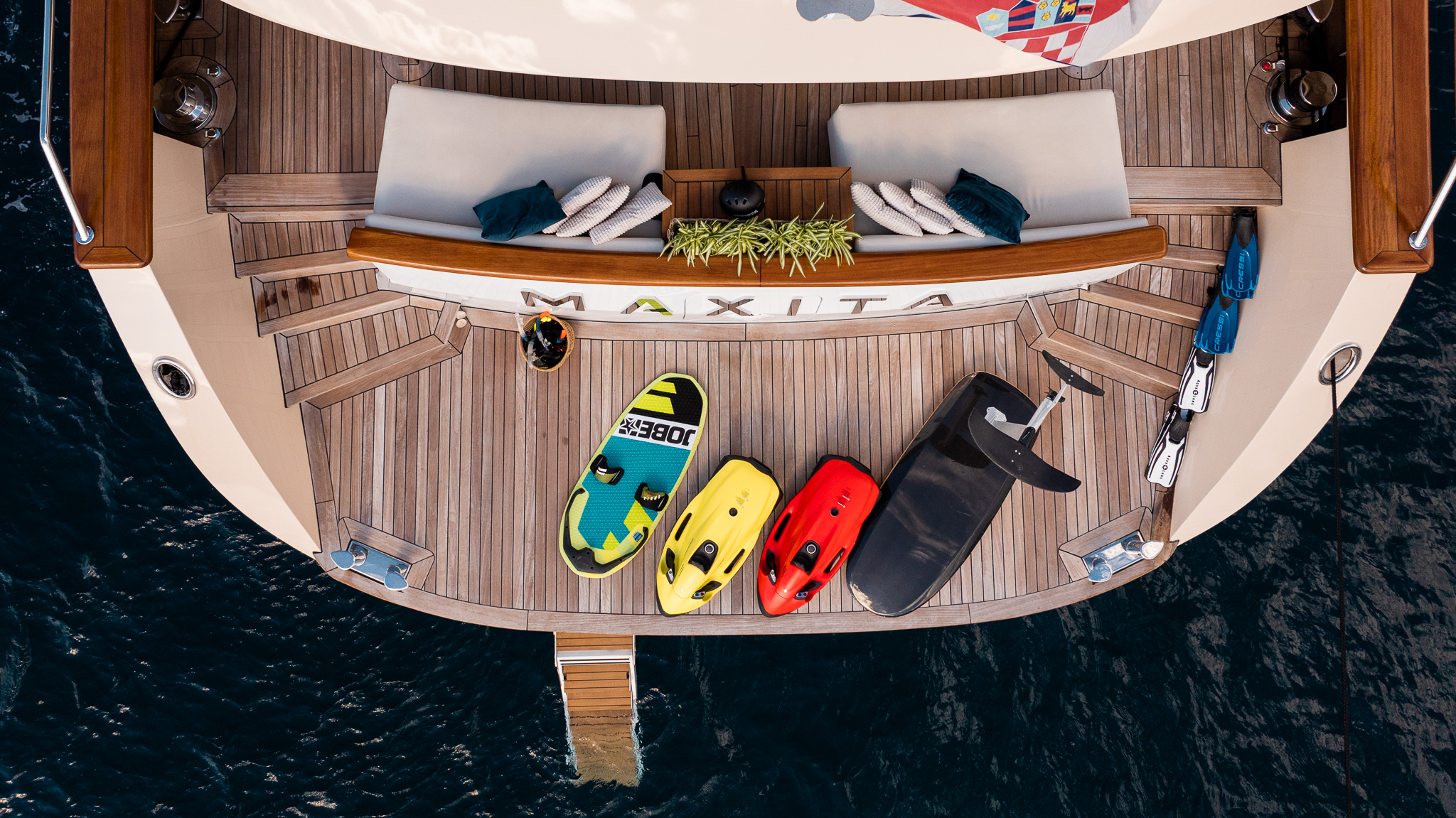 MAXITA Aerial view of the stern of a yacht named MAXITA, with two lounge chairs, towels, plants, and three colorful water sports boards on the deck above calm, dark water.