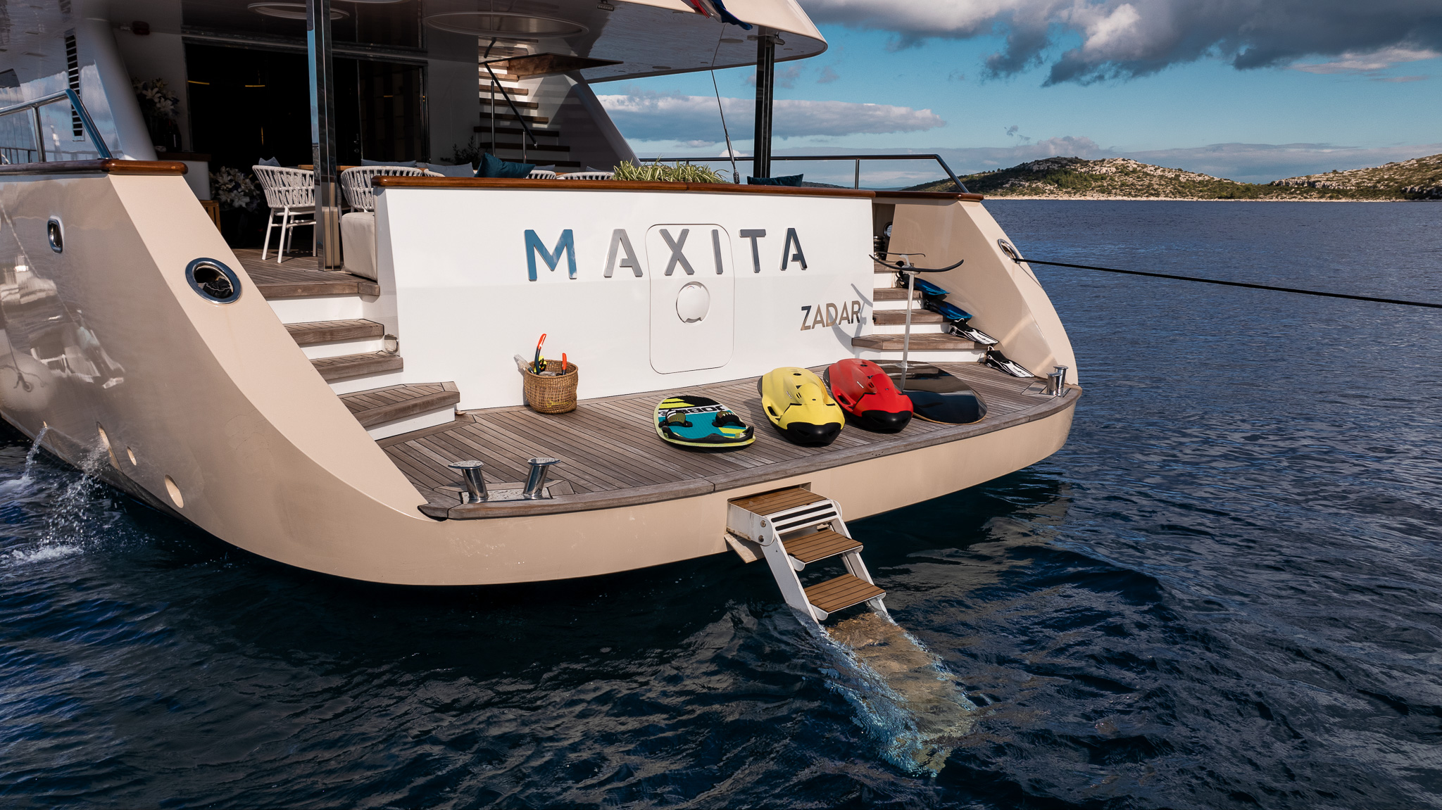 MAXITA The back of a yacht named MAXITA is anchored on calm blue water, with a swim platform, a small ladder leading to the sea, water sports gear, and a basket with drinks placed on the deck. Islands are visible in the distance.