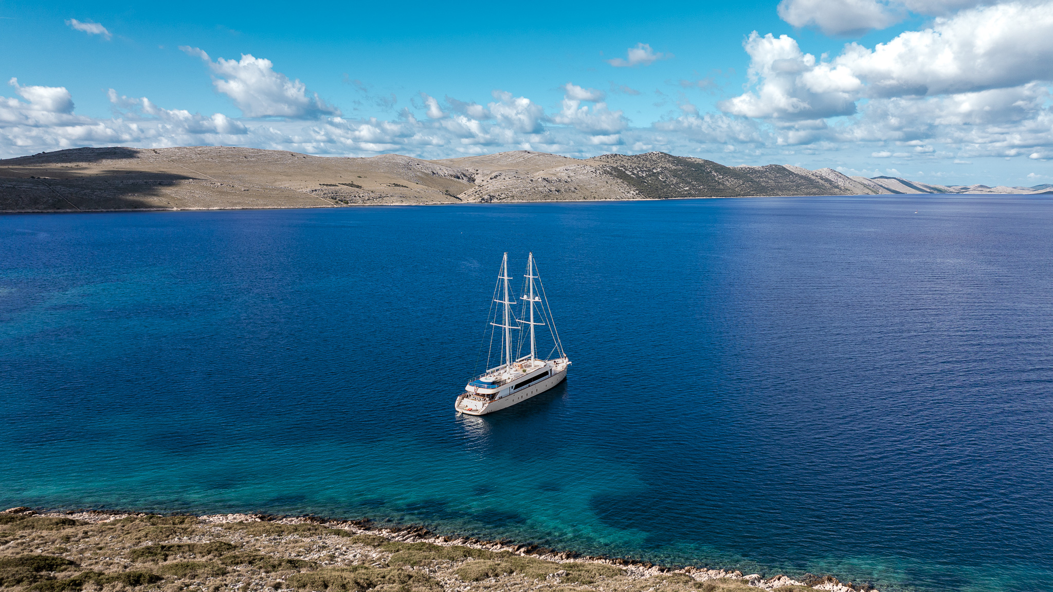 MAXITA A large sailboat with three masts floats on calm, deep blue water near a rocky shoreline. Rolling hills and scattered clouds appear in the background under a bright blue sky.